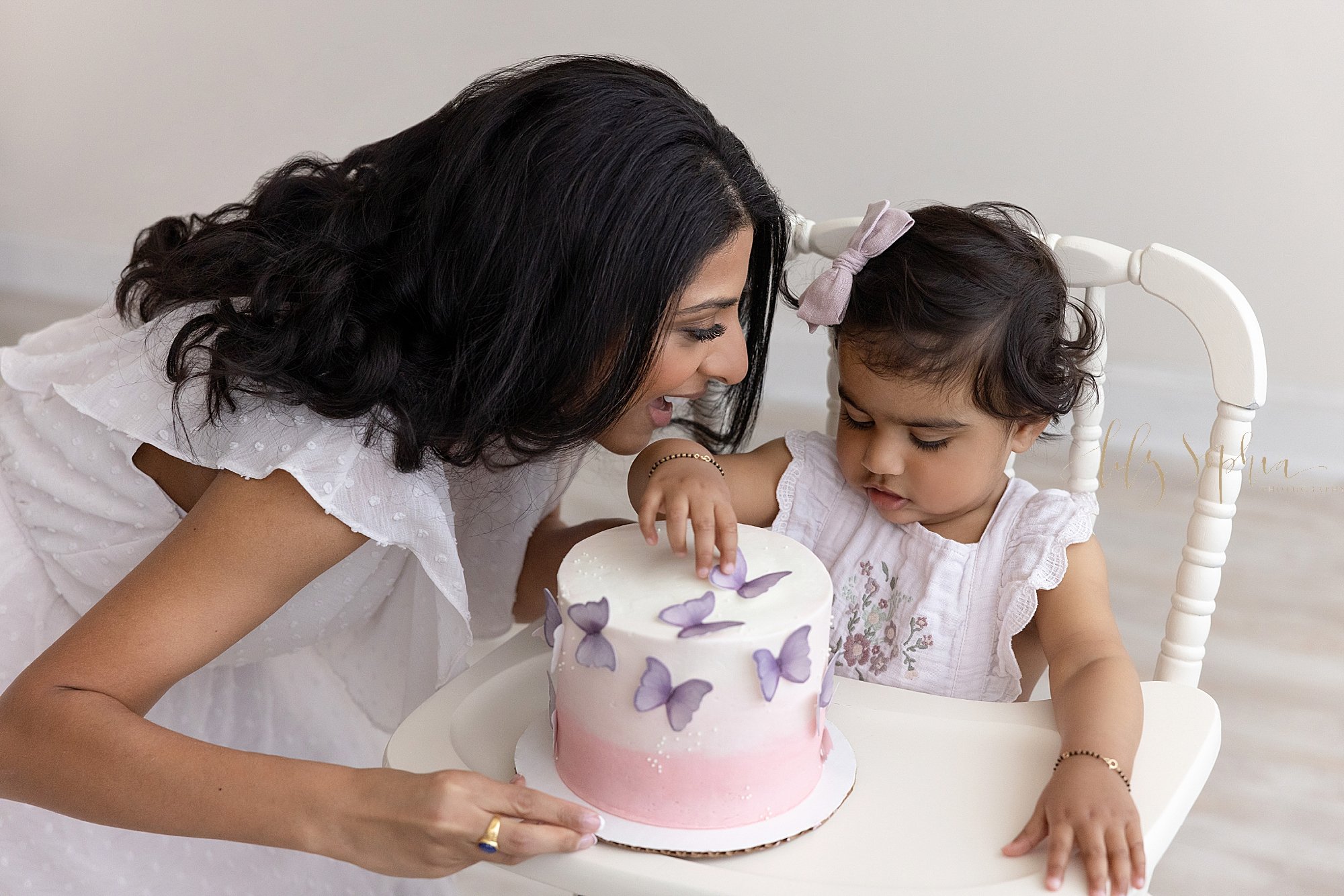  First birthday celebration with an overhead picture of a one year old girl sitting in an antique highchair as she touches the butterflies on her cake and her mother looks on taken in a natural light studio near Old Fourth Ward in Atlanta. 