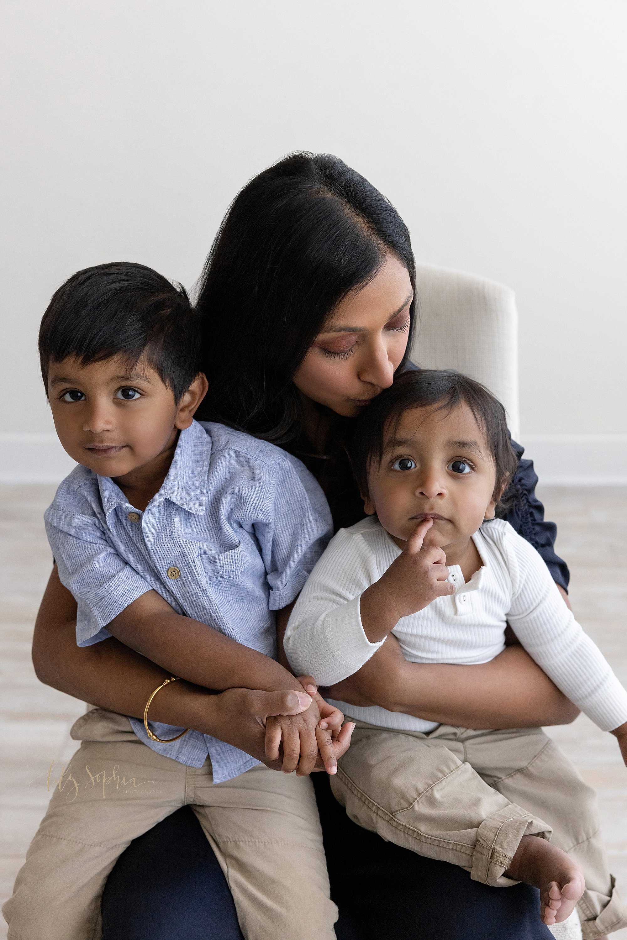  Family portrait of an Indian mother sitting on a chair with her one year old son on her left knee and her older son on her right knee as she bens her head and kisses her son celebrating his first birthday on his head taken in a natural light studio 