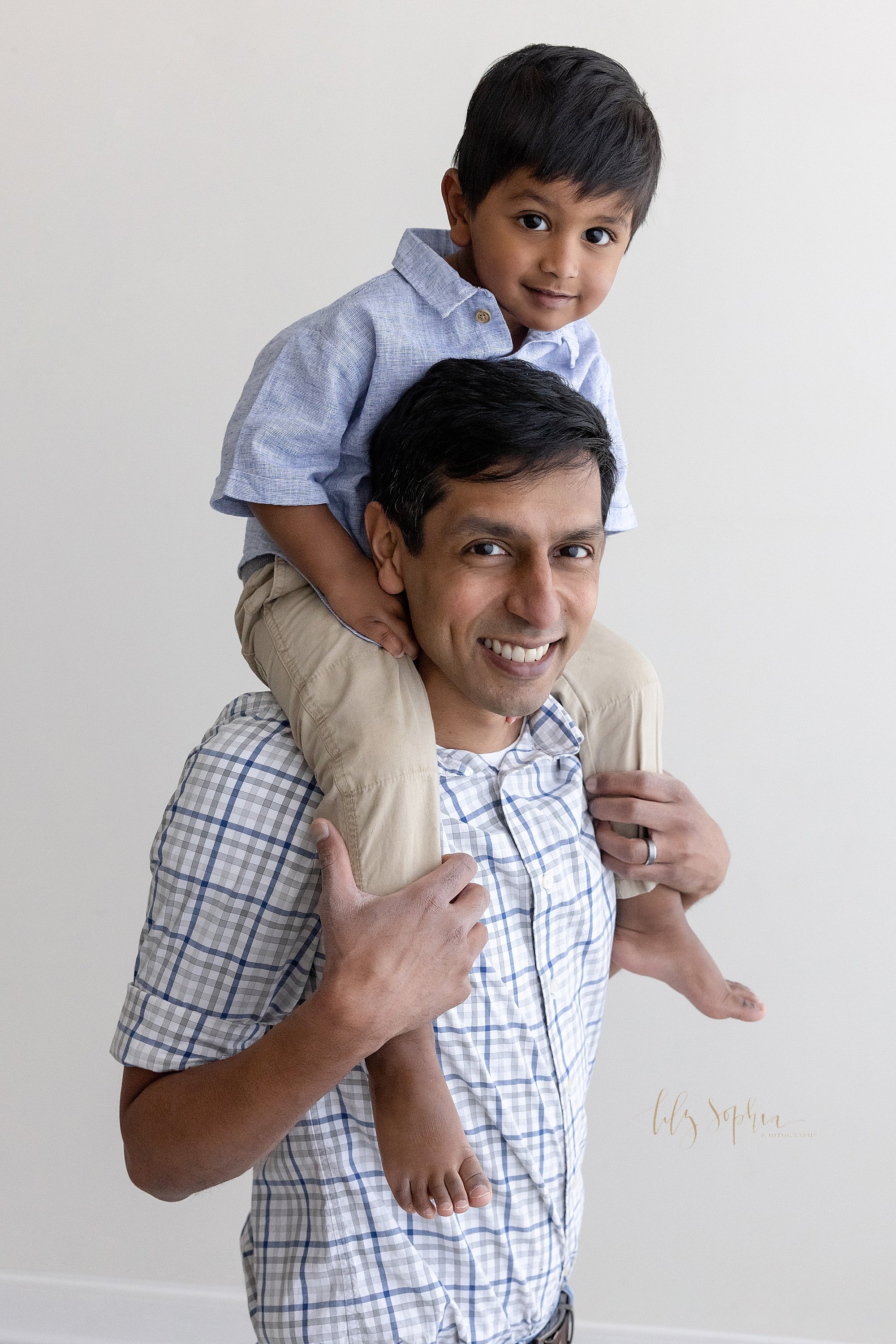  Father and son family picture with the young Indian  son sitting on his father’s shoulders as he stands in a studio near Buckhead in Atlanta that uses natural light. 