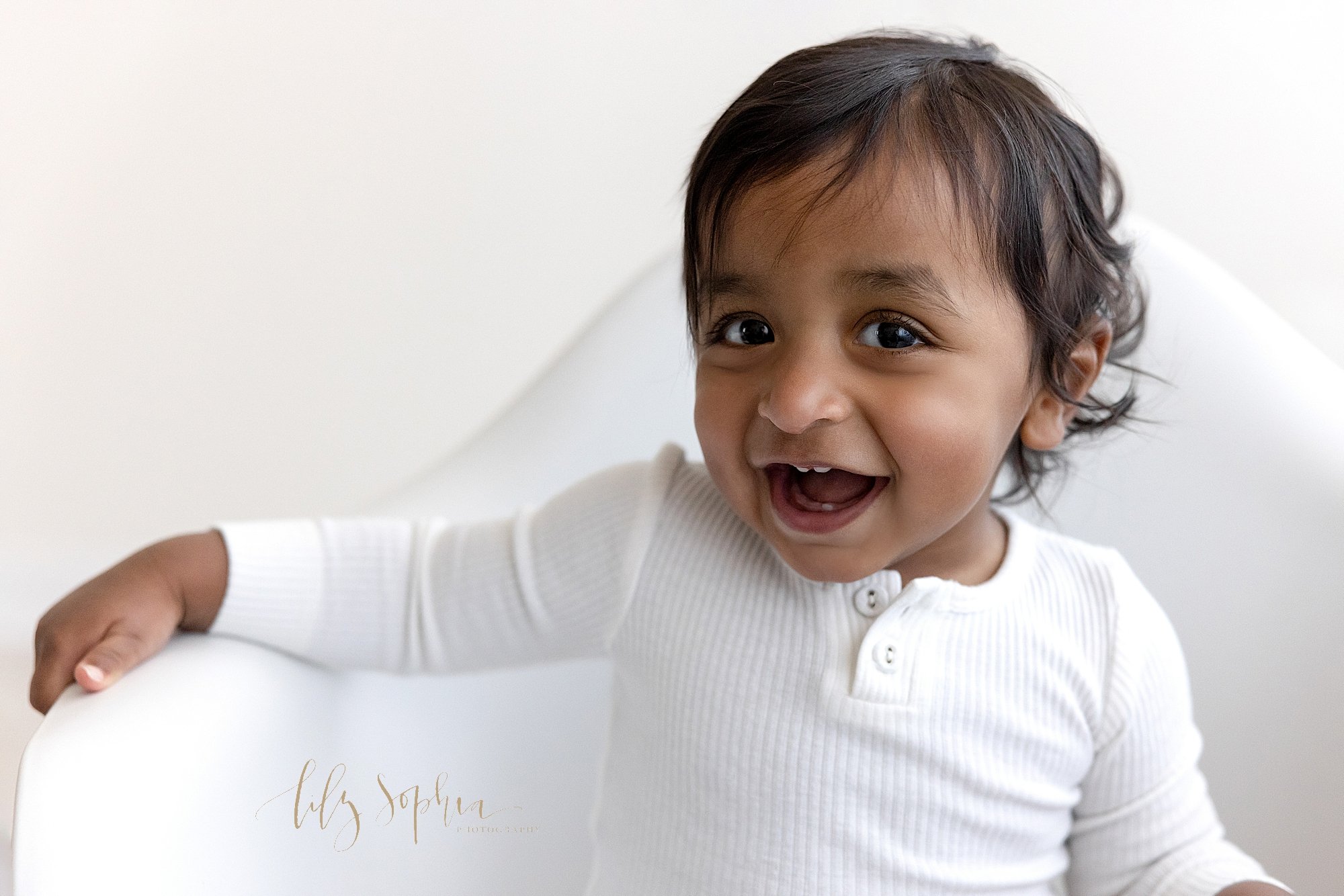  First birthday picture of a one year old Indian boy as he sits in a white molded chair laughing and showing his baby teeth taken near Decatur in Atlanta in a studio using natural light. 