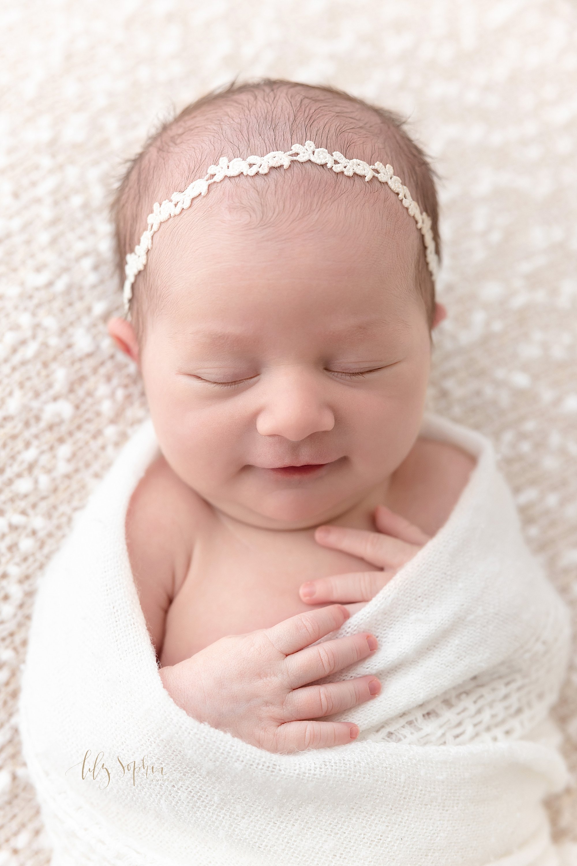  Close-up newborn portrait of an infant baby girl lying on her back wrapped in a soft white knitted  blanket as she grins in her sleep taken near Sandy Springs in Atlanta in a natural light studio. 