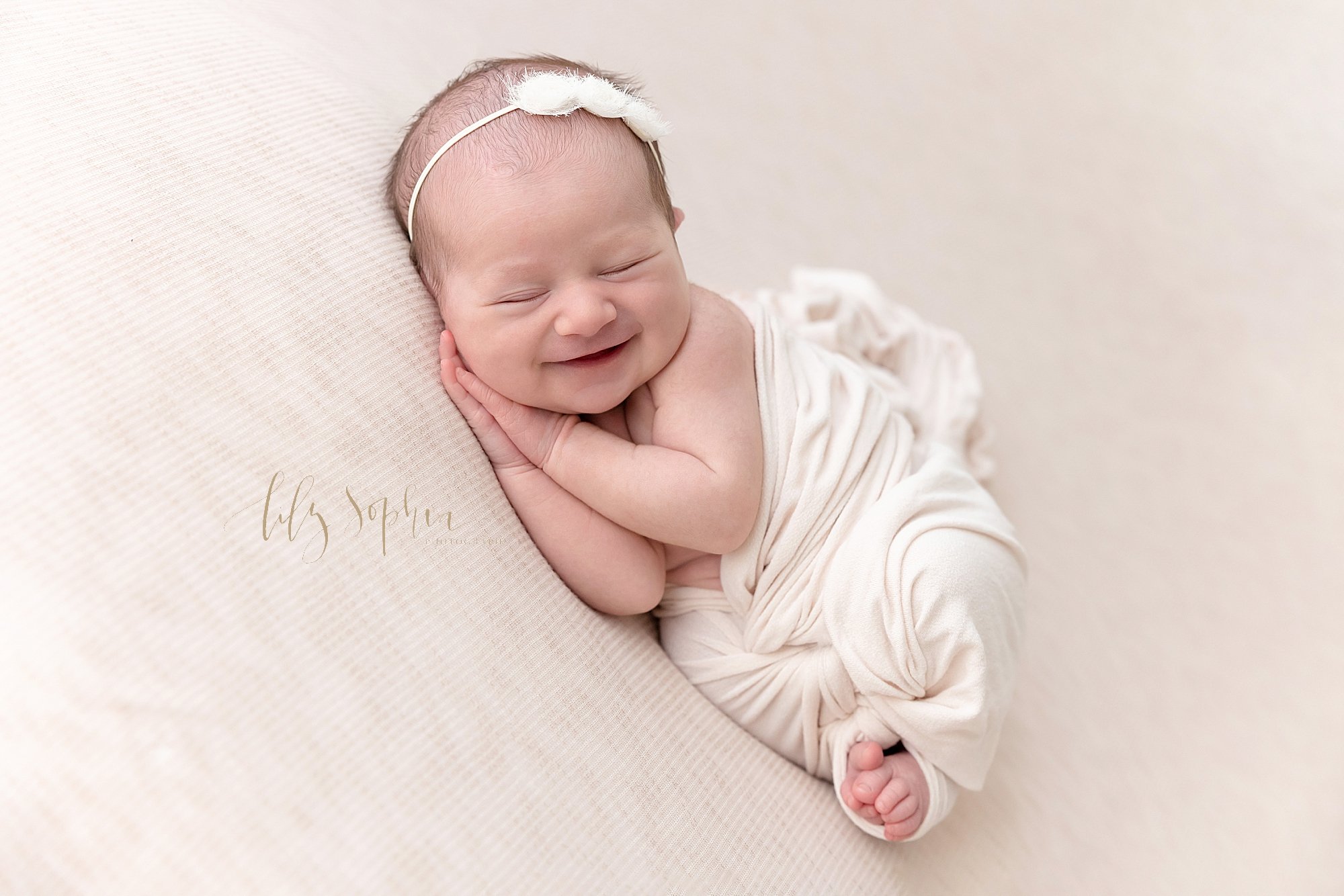  Newborn photo session with a smiling baby girl wearing a rose headband on her head while lying on her right side with her hands under her right cheek wrapped in a stretchy swaddle around her chest so her tiny toes peek out taken in natural light in 