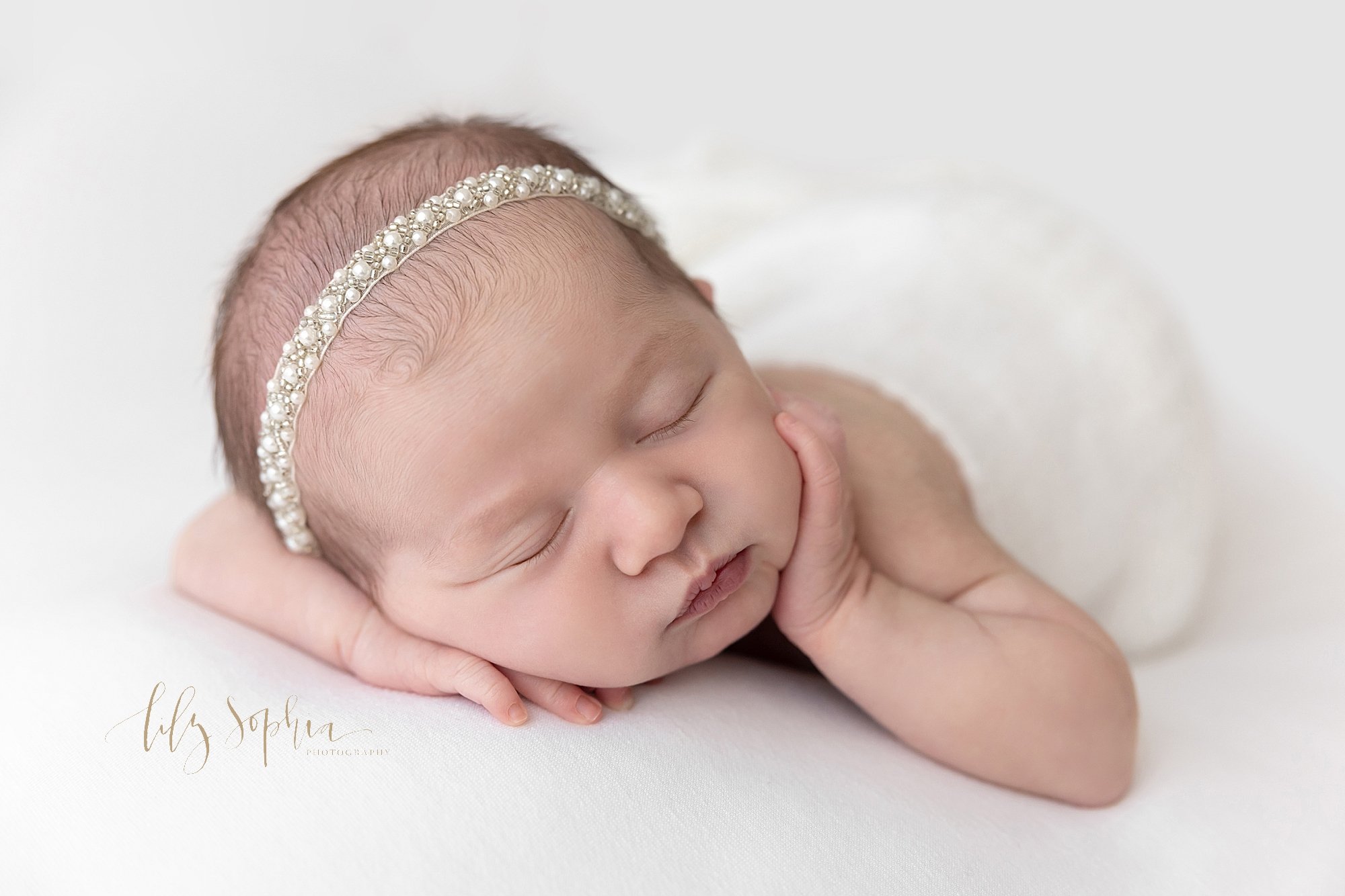  Newborn portrait of a baby girl as she lies on her stomach wearing a pearl headband on her head with her left hand on her cheek and her head resting on her right arm while draped with a soft white blanket taken in a studio near Ansley Park in Atlant