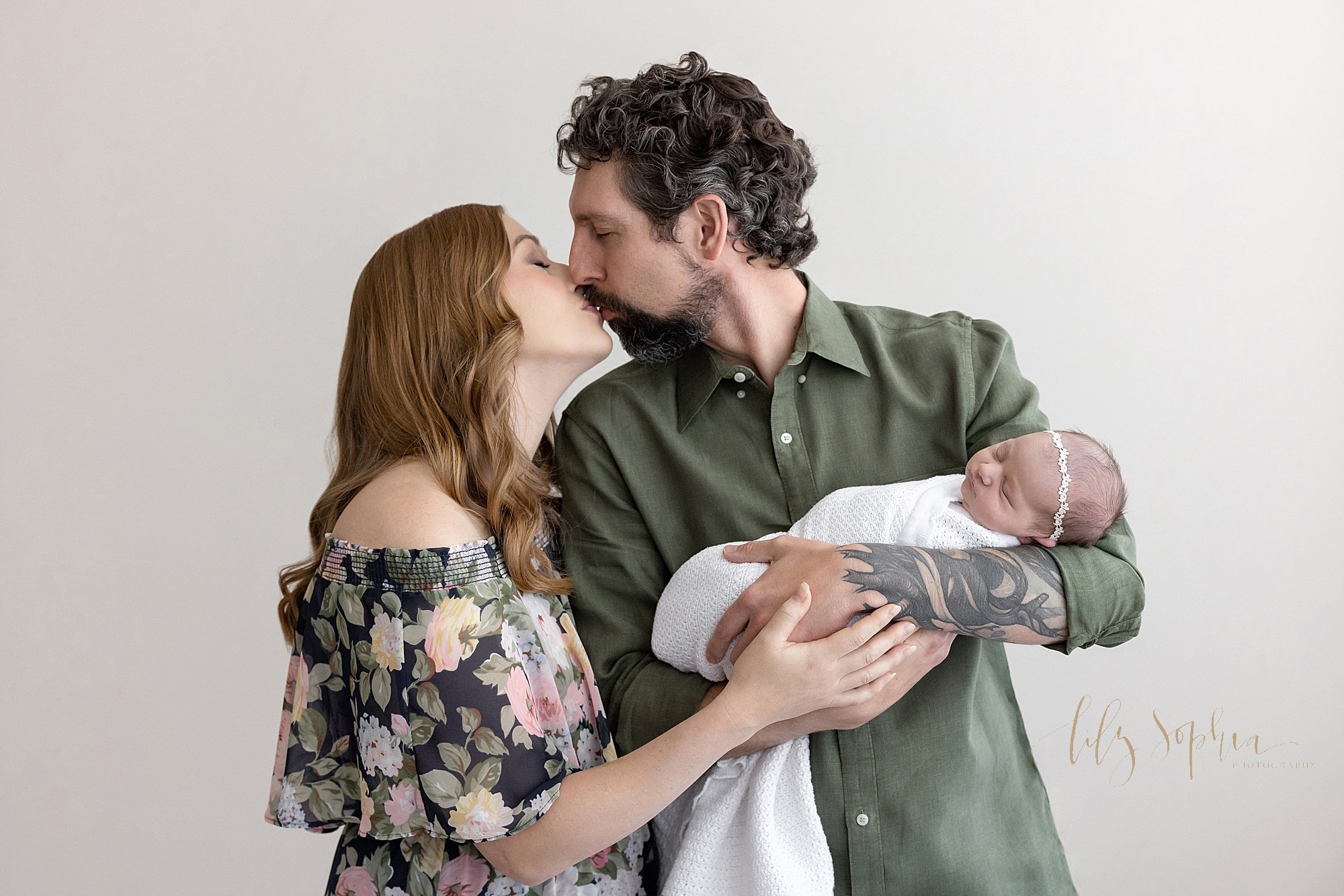  Family newborn photo of a father cradling his peacefully sleeping infant daughter in his arms as he looks over his right shoulder and kisses his wife who is standing next to him in the natural light photography studio located near Morningside in Atl