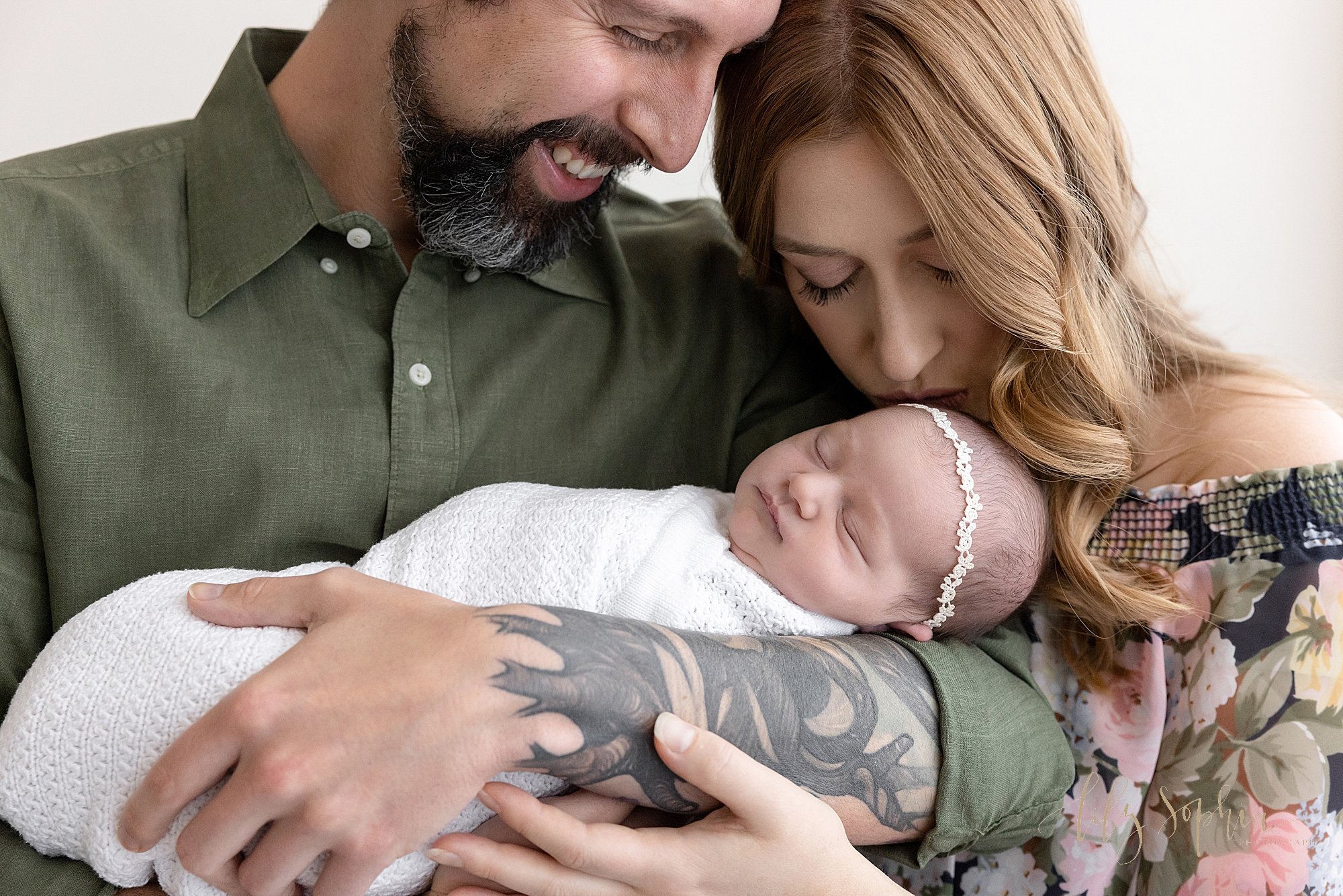  Family newborn photo of a father cradling his sleeping infant daughter in his arms as his wife stands to his left and bends her head to kiss her daughter’s head while her husband smiles proudly taken using natural light near Virginia Highlands in At