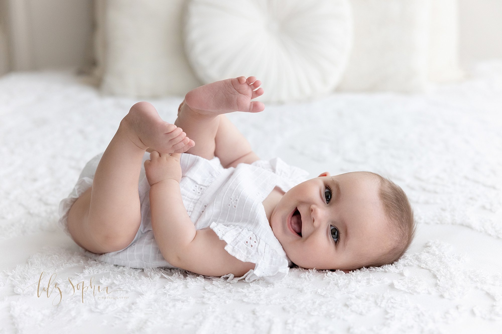  Milestone baby photo session of a smiling baby girl as she lies on her back with her feet up in the air on a bed in a natural light studio taken near Midtown in Atlanta, Georgia. 