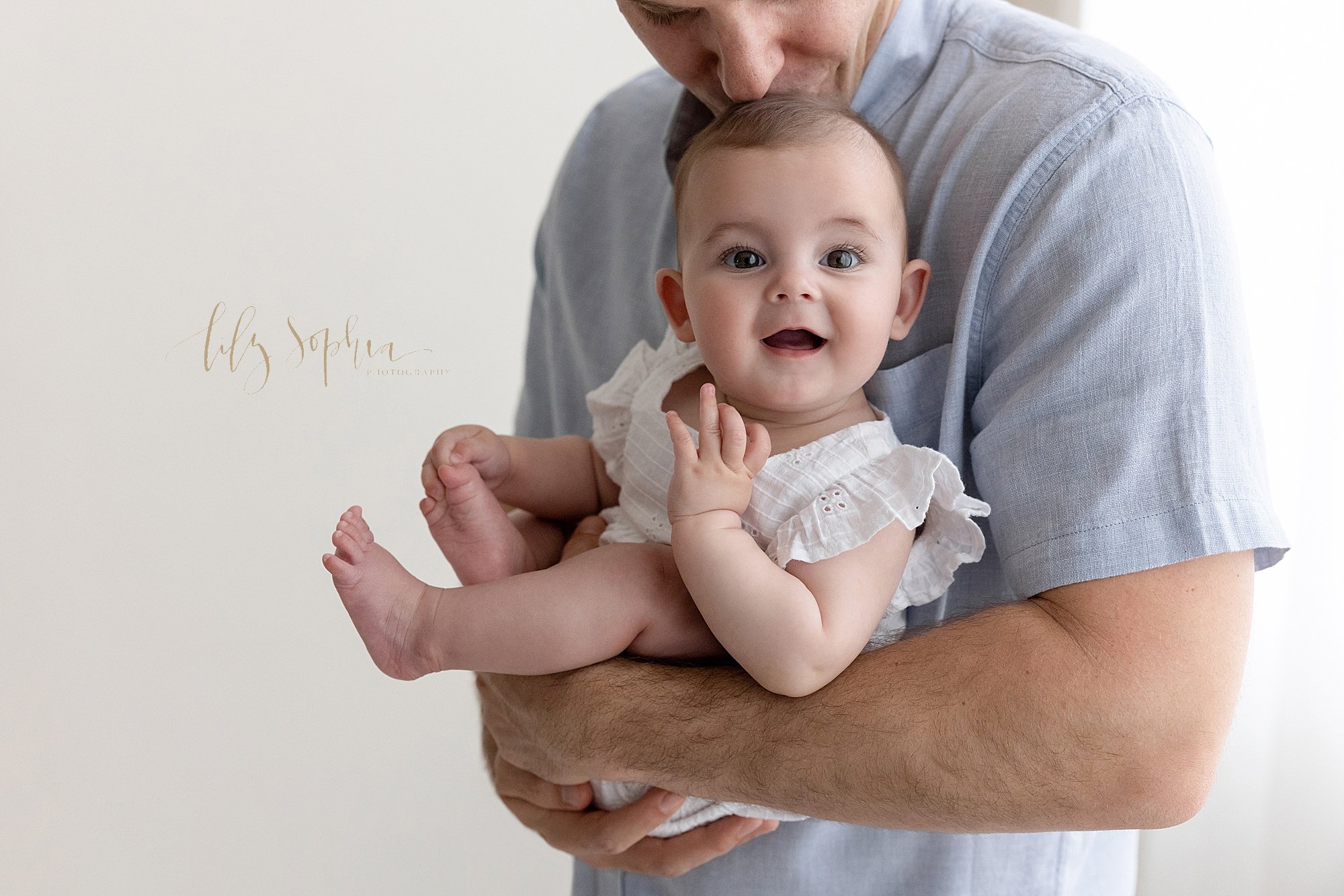  Family portrait of a father holding his baby girl in front of him as she plays with her toes with her right hand and dad kisses the top of her head while standing in front of a natural light window in a studio near Poncey Highlands in Atlanta. 