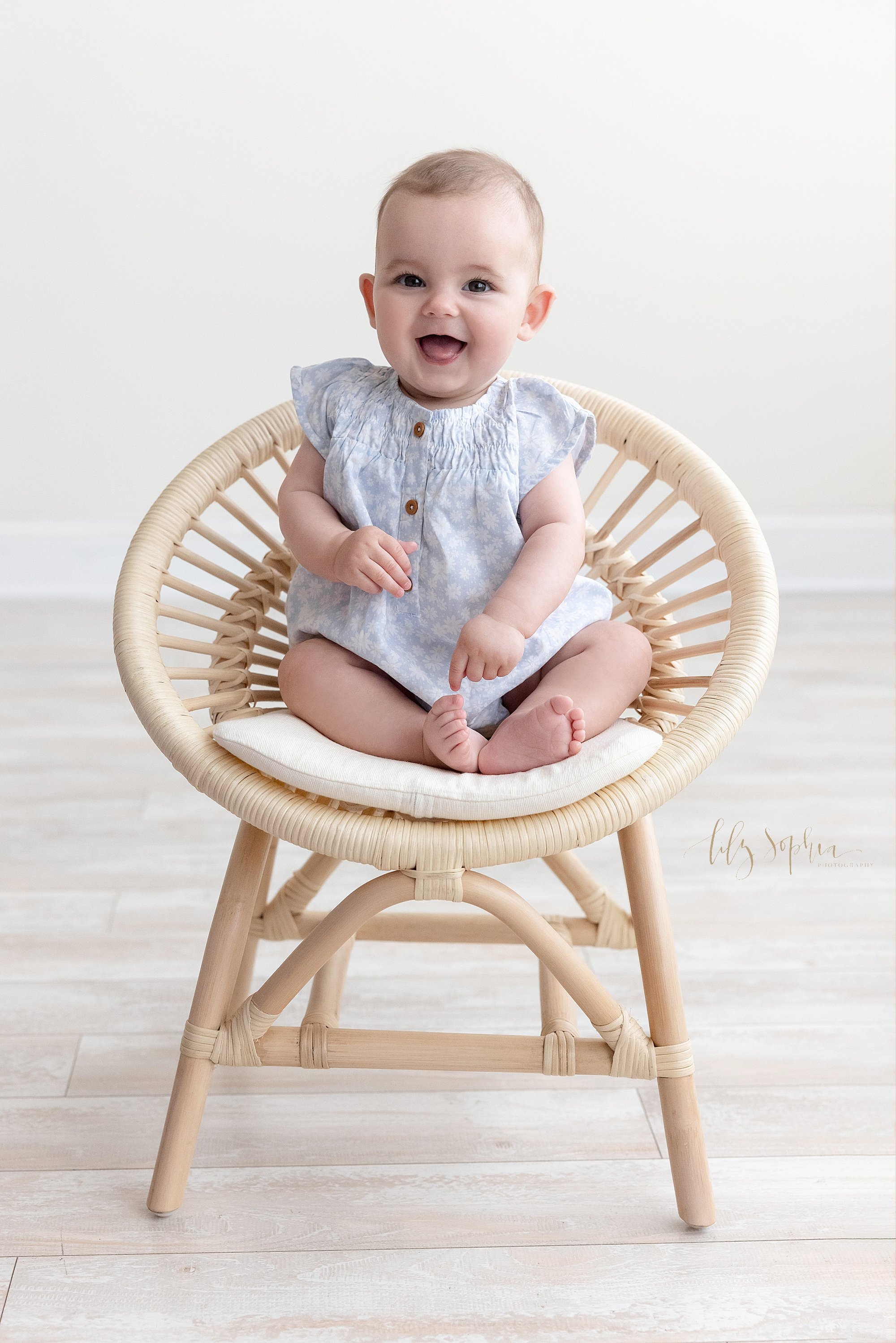  Sitting milestone portrait of a smiling baby girl as she sits in a rattan chair taken near Old Fourth Ward in Atlanta in a natural light studio. 