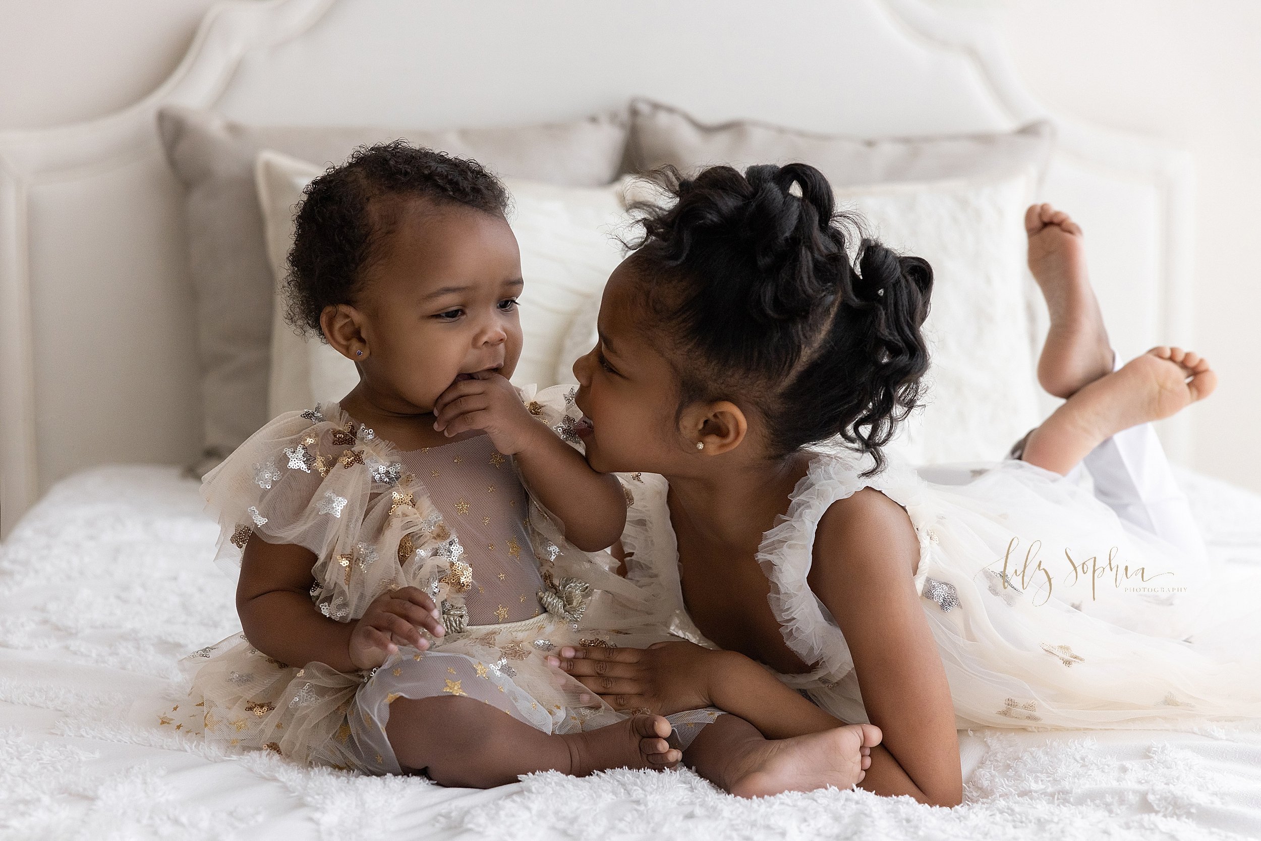  Sibling portrait of two African-American sisters as the older one lies on her stomach on a bed with her feet crossed behind her and her one year old sister sits next to her and the two of them talk taken near Decatur in Atlanta in a natural light st