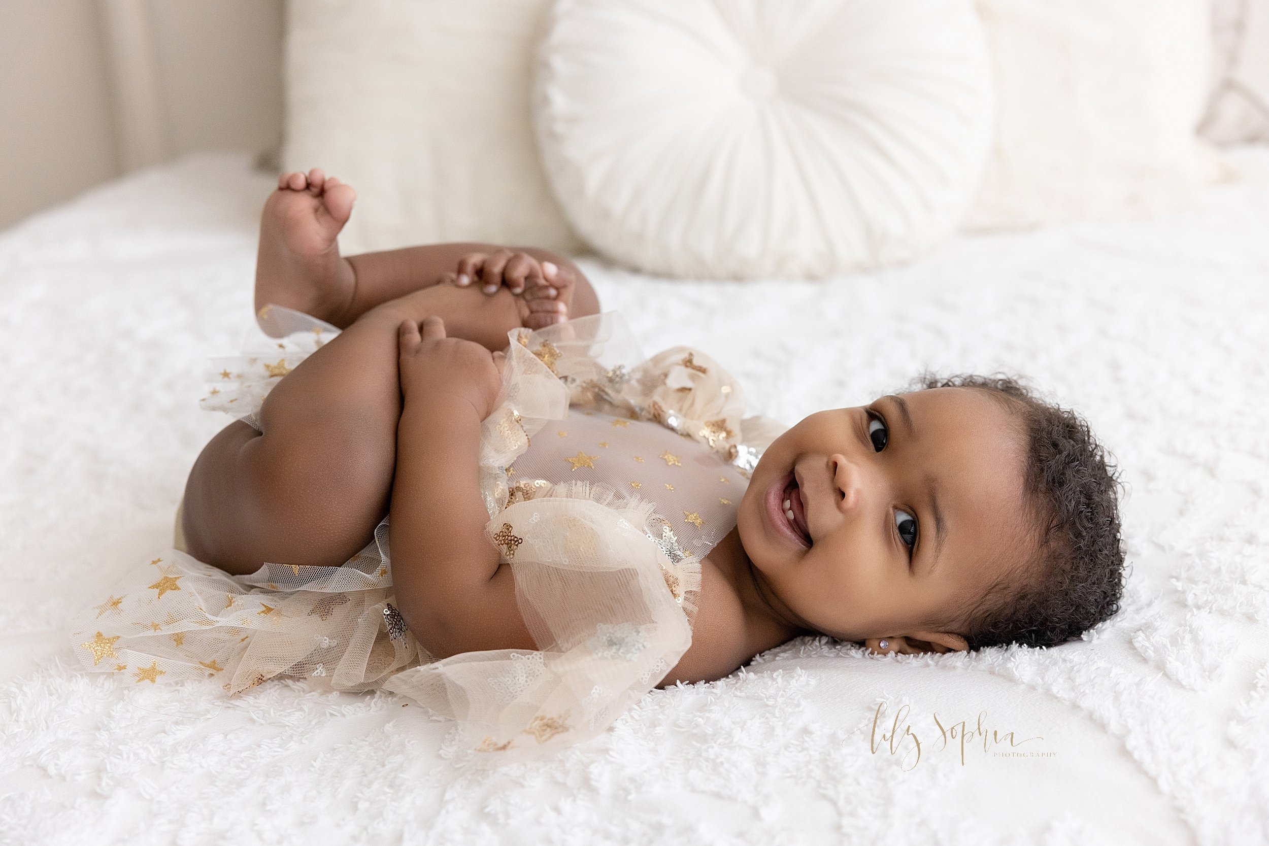  First birthday picture of an African-American girl lying on her back playing with her feet on a bed while smiling to show her little baby teeth taken in a studio that uses natural light near Roswell in Atlanta. 