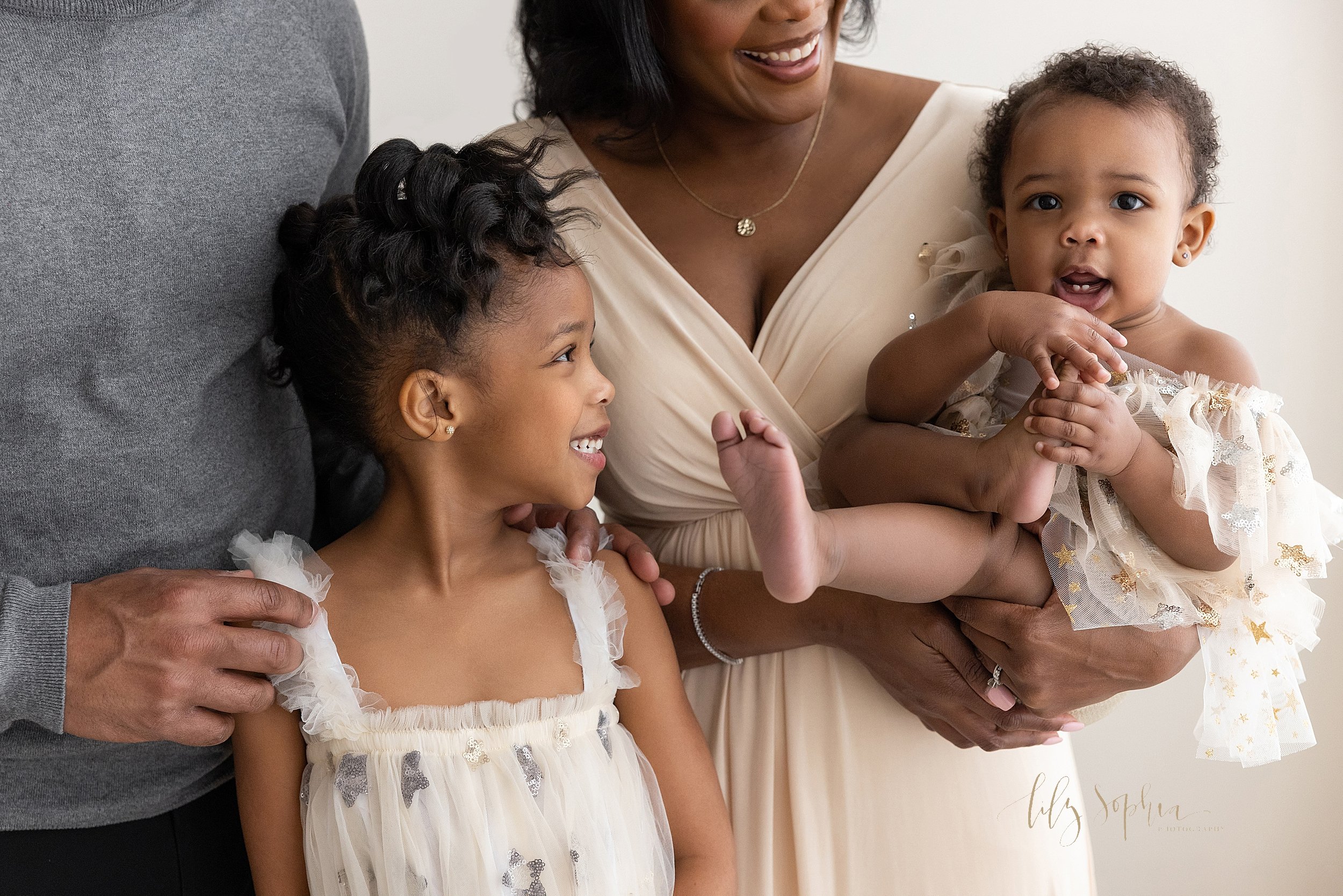  Family portrait of an African-American mother holding her one year old daughter in her right arm as she crosses her right leg over her left knee to play with her toes as her husband stands to her right side with their young daughter standing between