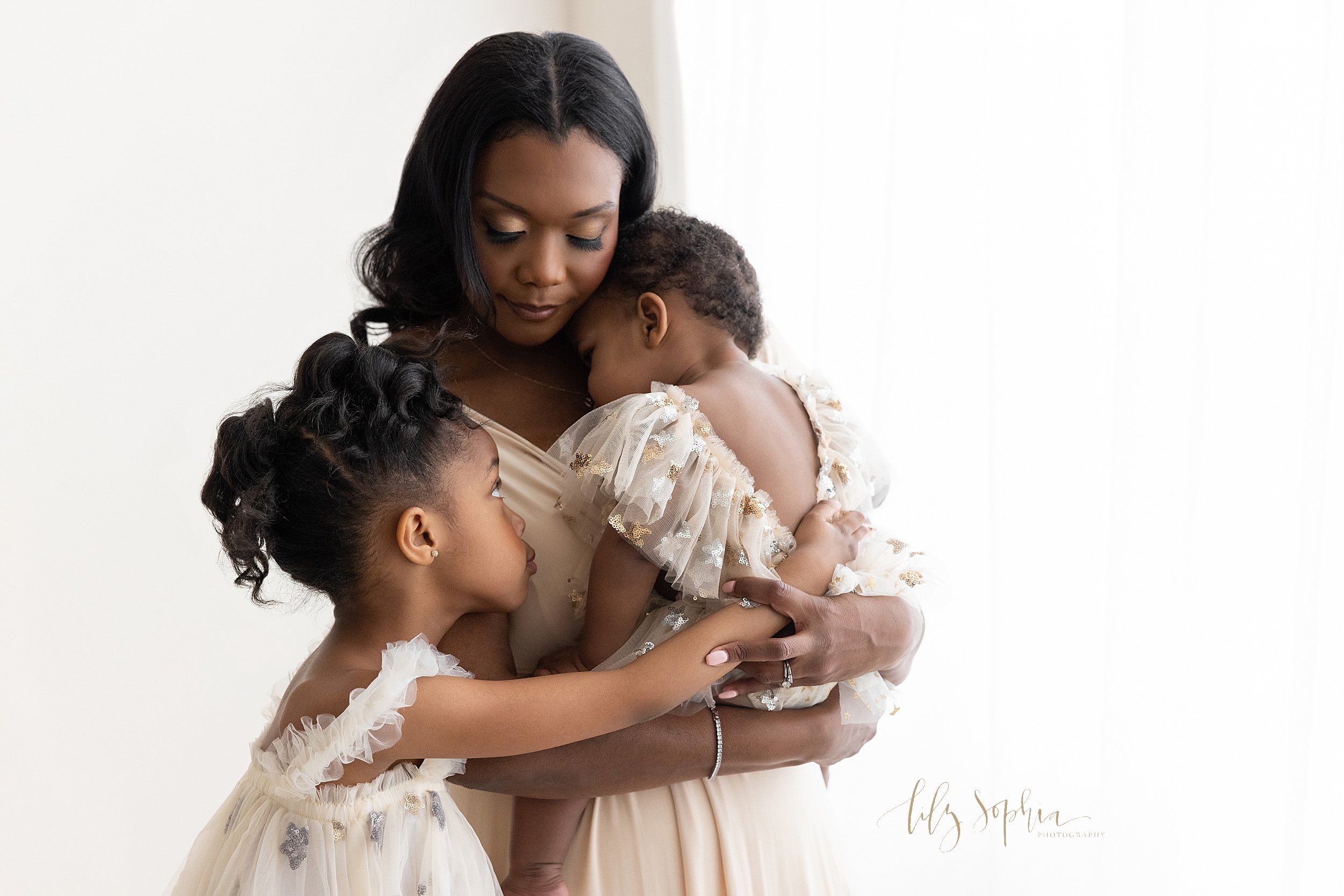  Family portrait of an African-American mother as she holds her one year old in her arms and her younger daughter stands to her mother’s right side and places her hand on her sister’s back taken in front of a window streaming natural light in a studi
