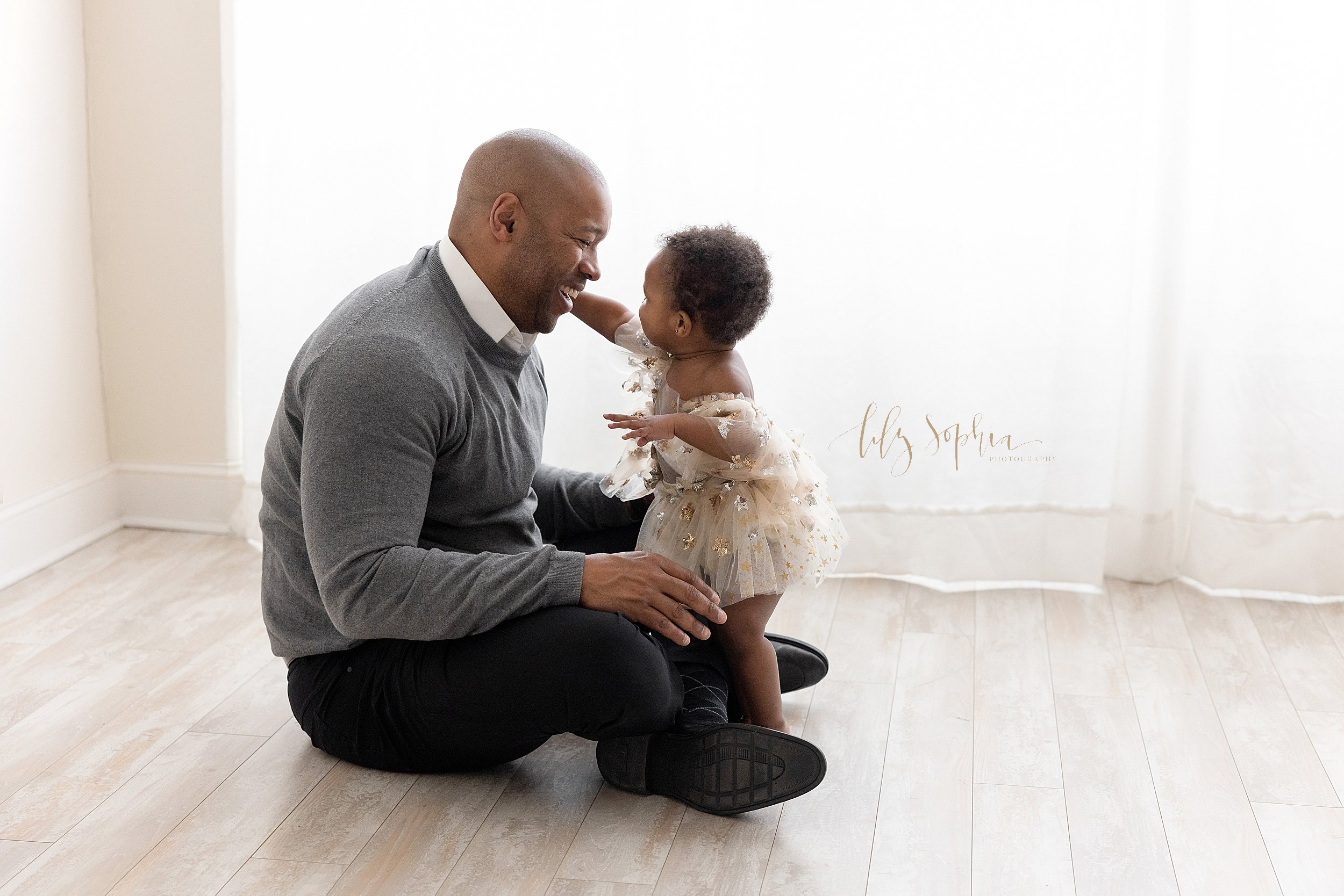  Family photo of an African-American father sitting on the floor of a natural light studio as his one year old daughter stands in front of him and reaches her right hand out to give him a hug taken near Oakhurst in Atlanta. 