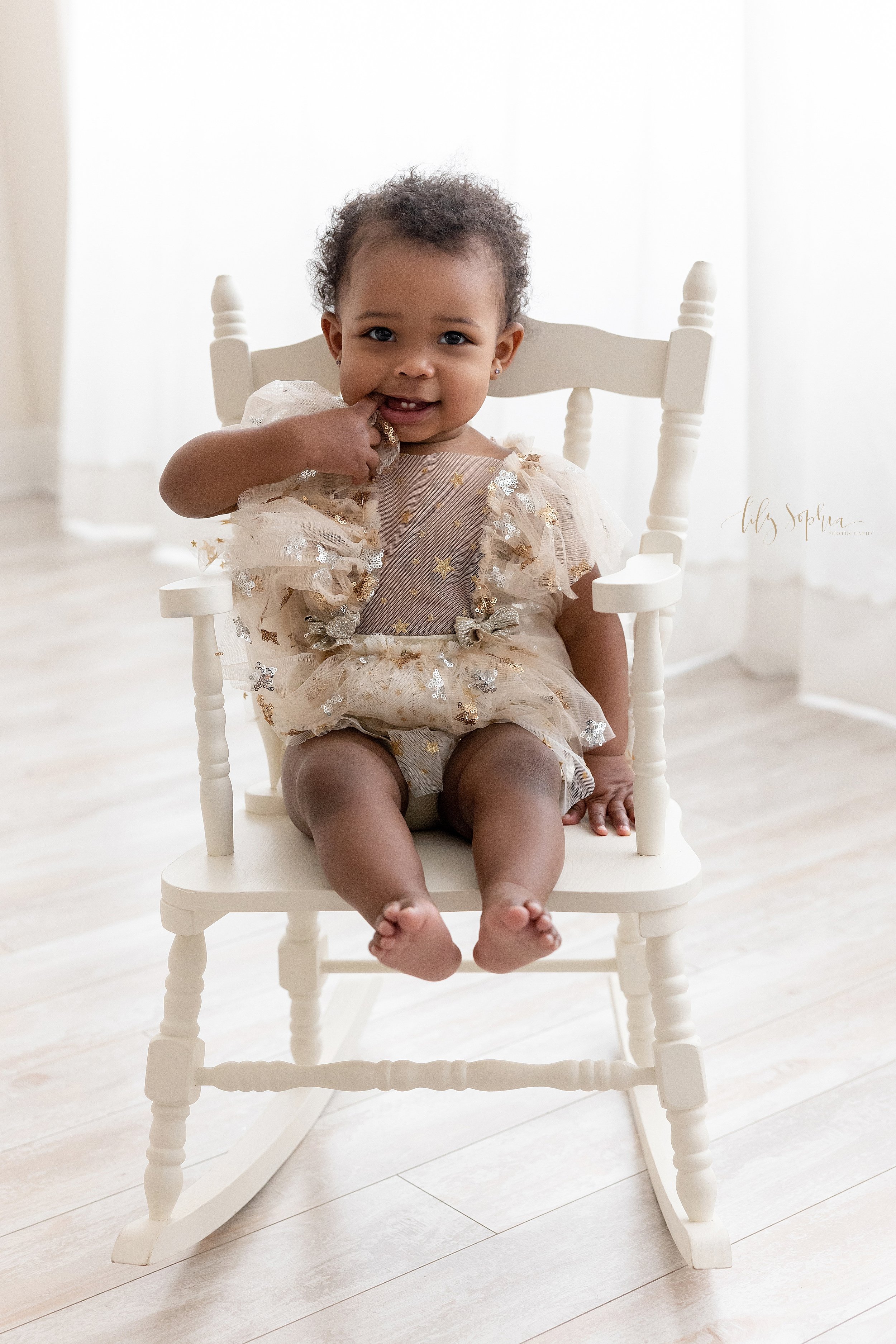  First birthday portrait of a one year old African-American baby girl as she sits in a rocking chair in front of a window streaming natural light and chews on her right finger taken in a photography studio in Ponce City Market. 