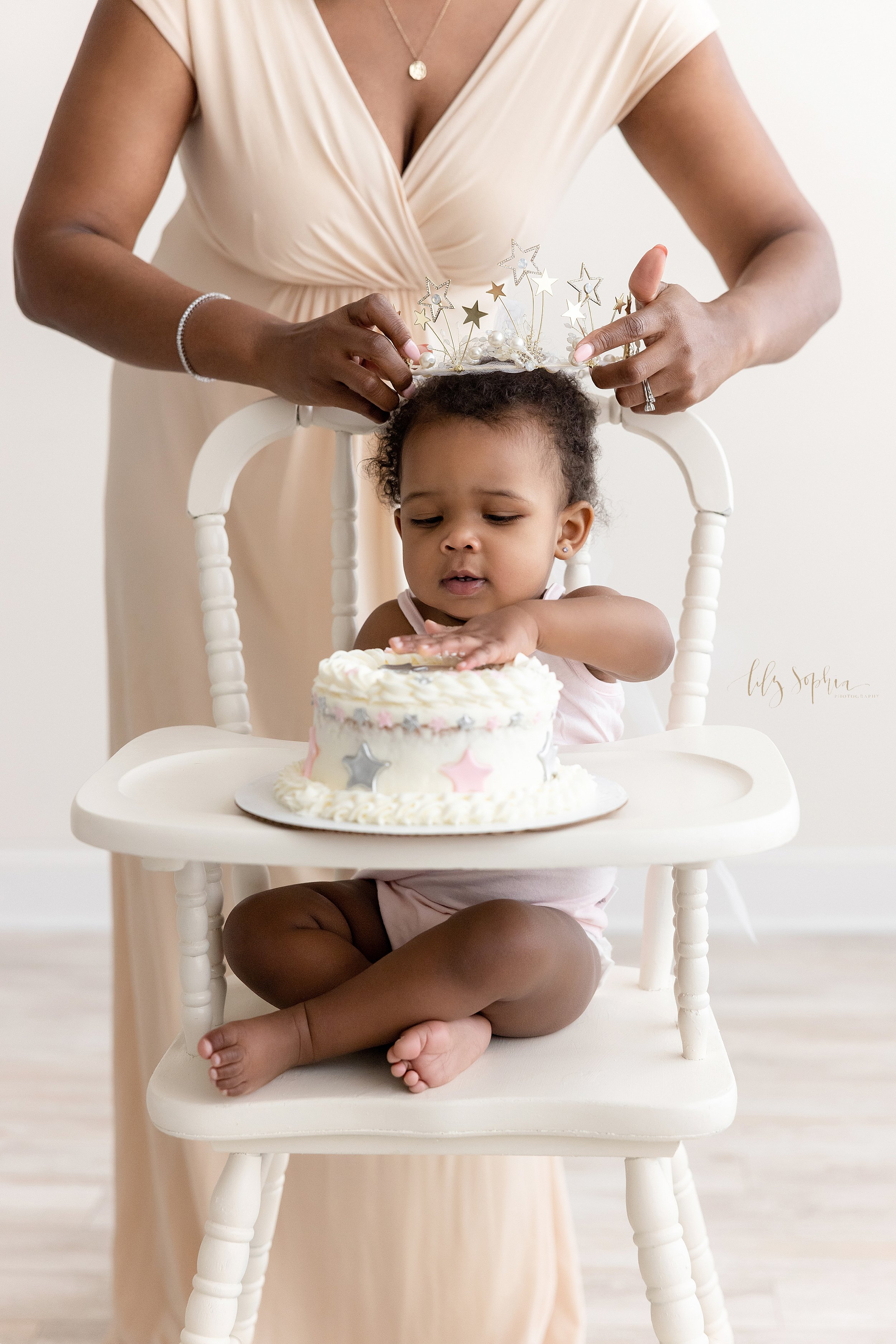  Smash cake photo shoot of a black one year old baby girl as she sits in a highchair with her first birthday cake on the tray and her mom stands behind her to put a crown on her head taken in a natural light photography studio near Kirkwood in Atlant