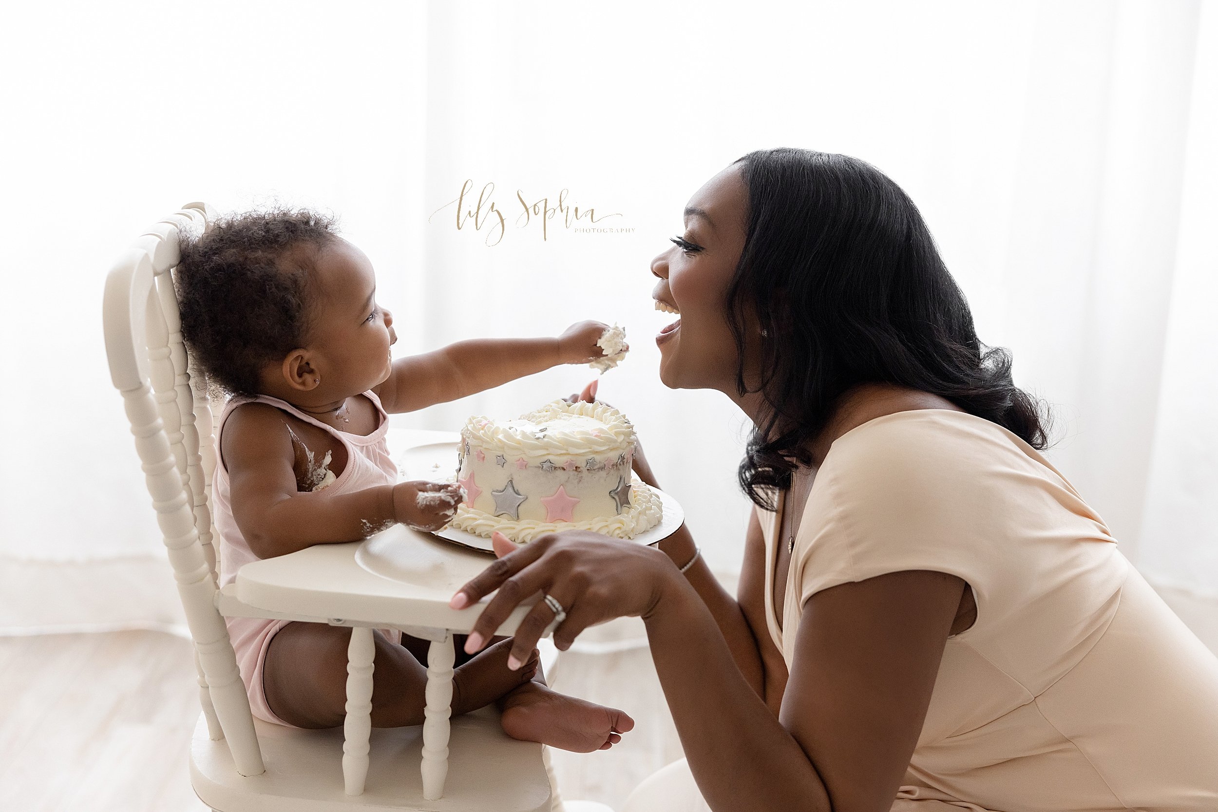  Smash cake photo session celebrating the first birthday of a black baby girl as her mom kneels in front of an antique high chair while the baby girl reaches to give a bite of her cake to her mother taken in front of a window streaming natural light 