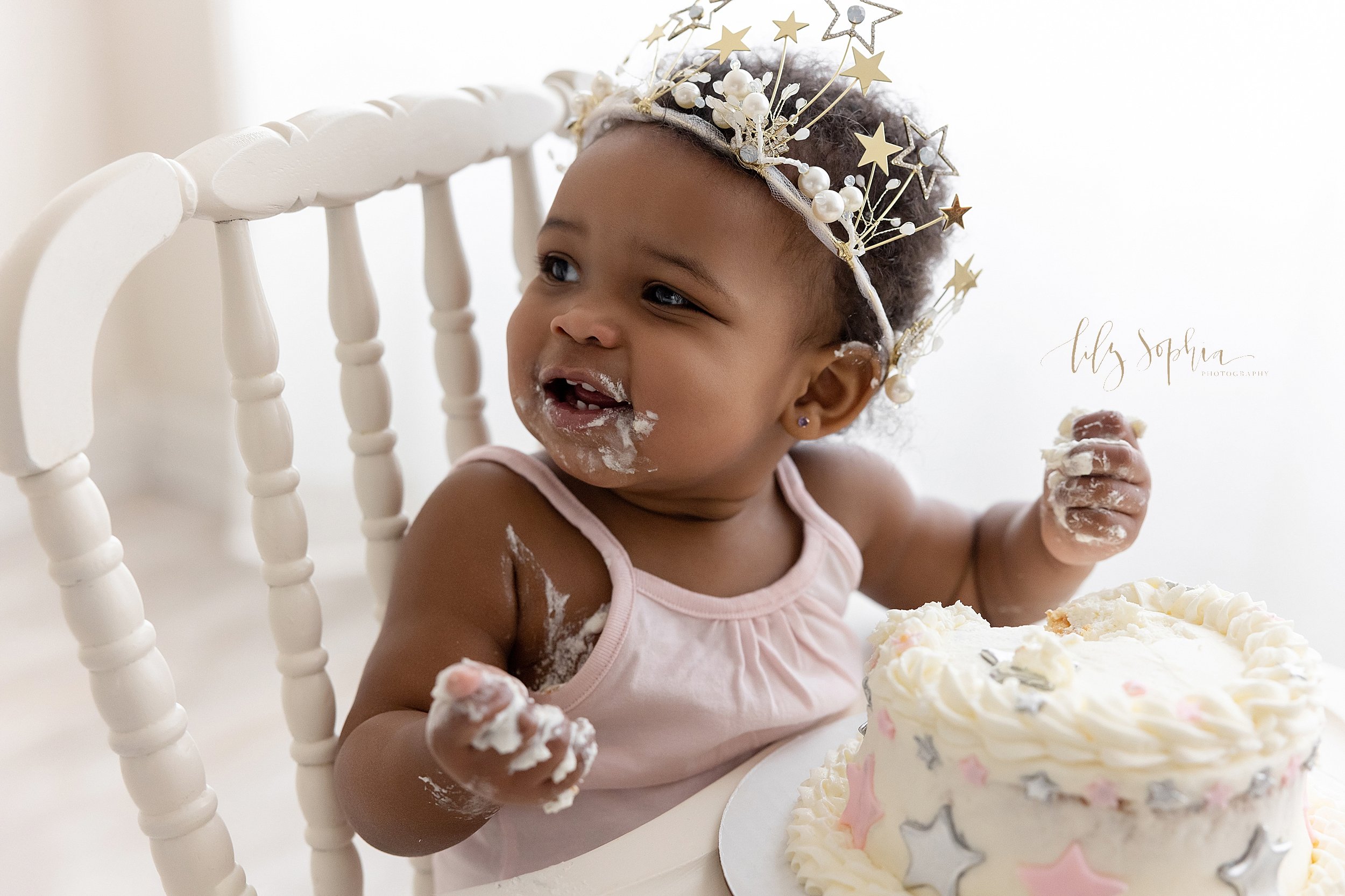  First birthday smash cake photo session with a one year old African-American girl wearing a sparkly crown on her head looking over her right shoulder as she sits in an antique high chair with icing covering her hands and her mouth taken in a studio 