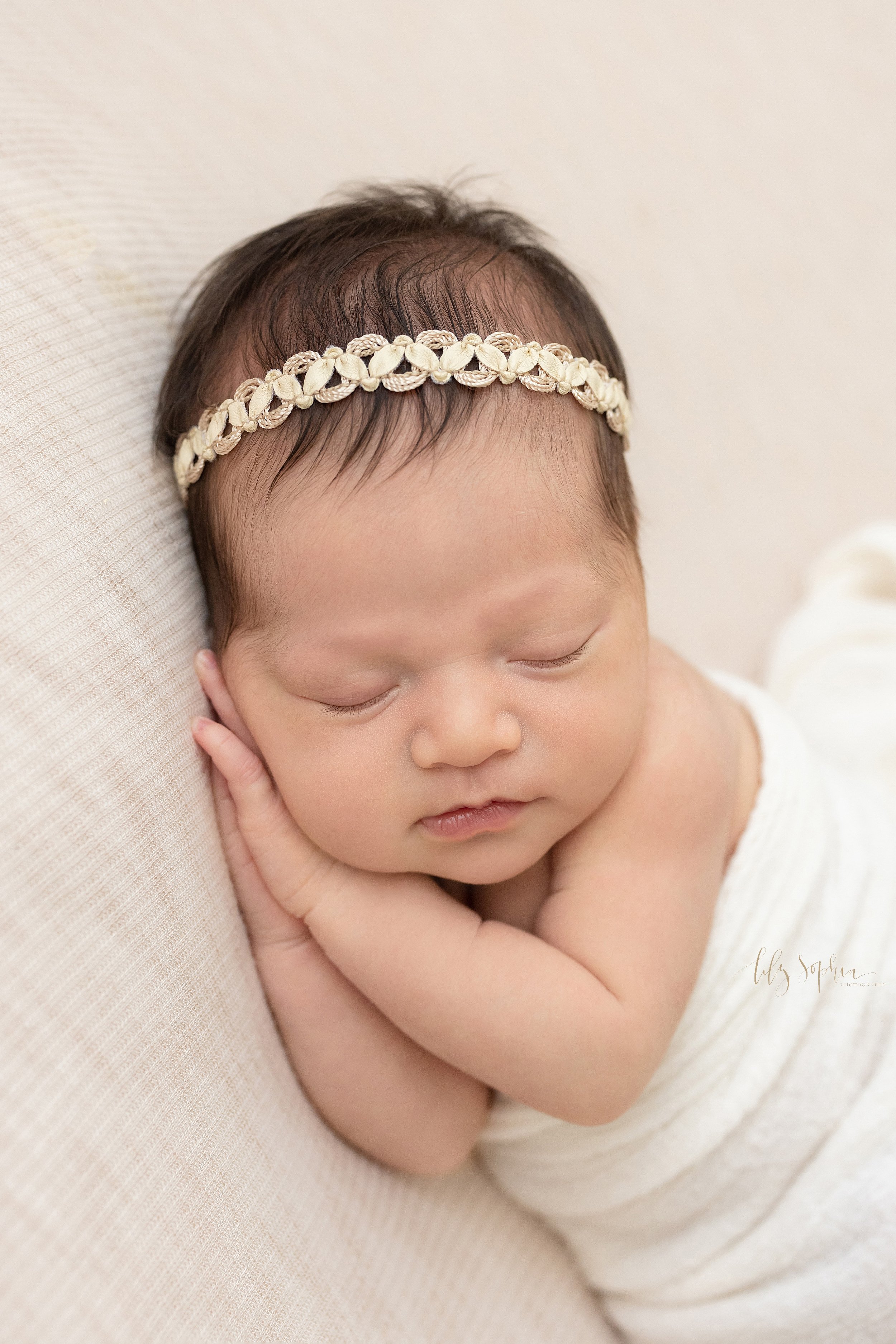  Close-up newborn portrait of a peacefully sleeping newborn baby girl with her hands under her right cheek and draped with a soft white knitted blanket taken in a natural light studio near Roswell in Atlanta, Georgia. 