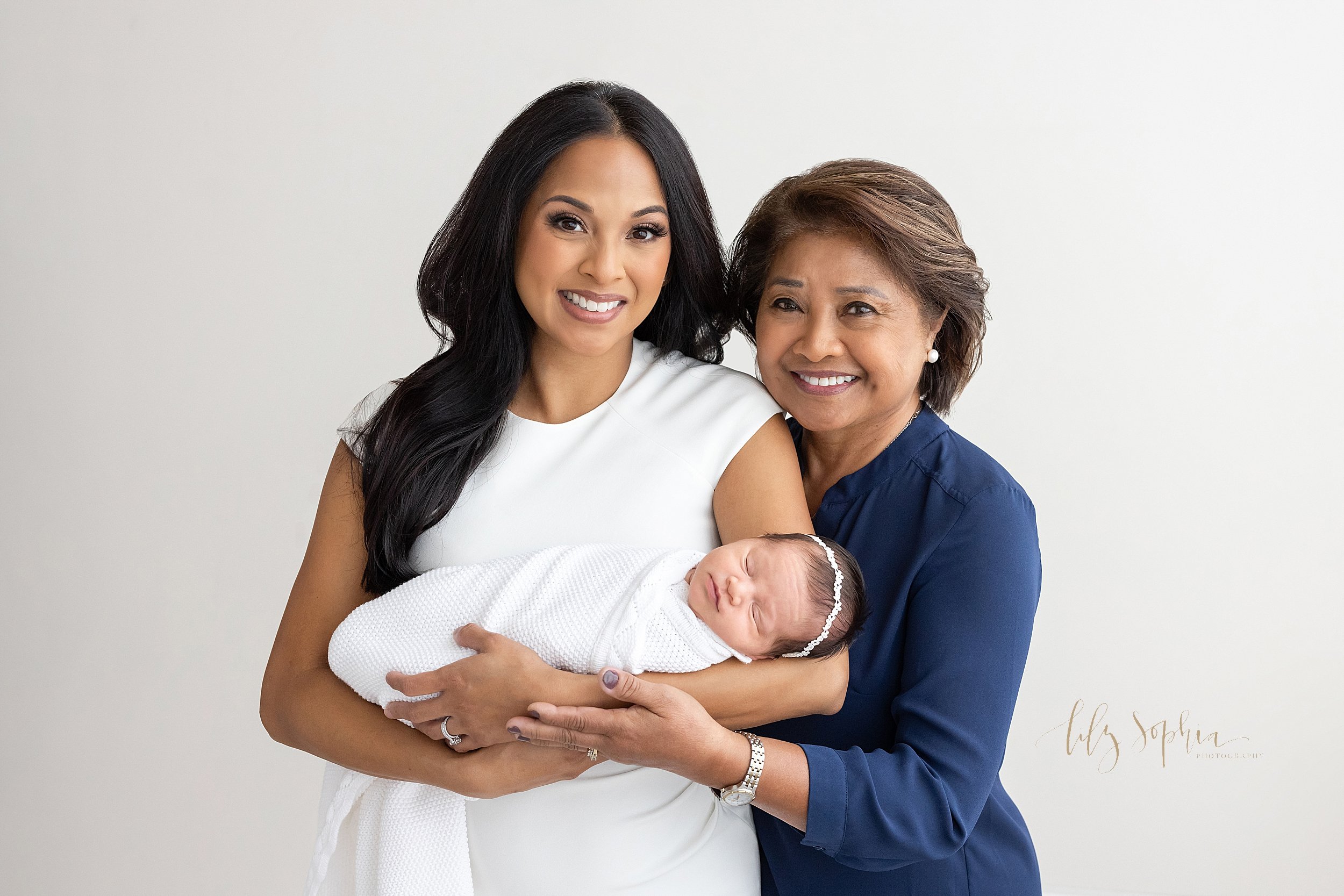  Multi-generational newborn photo session of a mother cradling  her peacefully sleeping newborn daughter in her arms as her mother stands to her left and places her left hand on her daughter’s to support her taken near Smyrna in Atlanta in a natural 