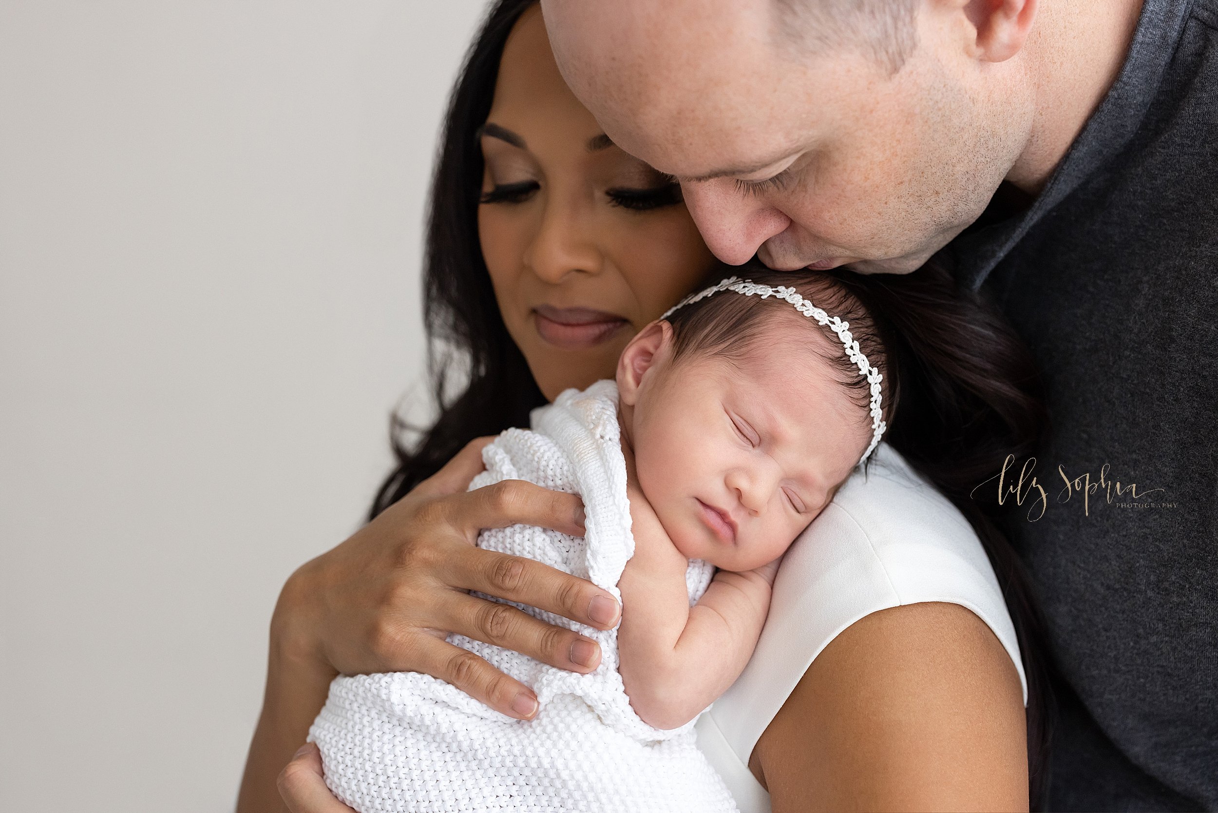  Family newborn picture of a mother holding her sleeping infant girl on her shoulder as her husband stands behind her and kisses his daughter’s head taken in a studio near Ansley Park in Atlanta that uses natural light. 