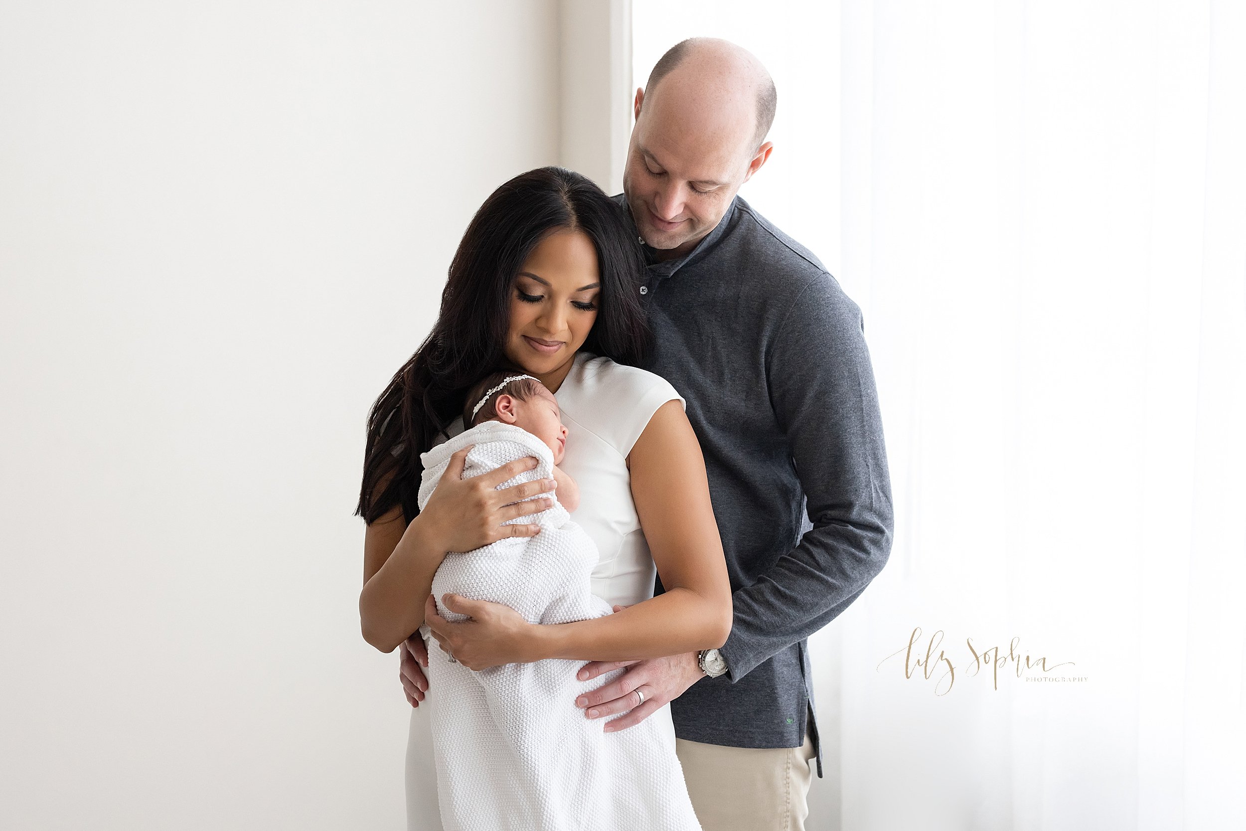  Newborn photo session of a mother holding her newborn baby girl against her chest as her husband stands behind her and looks down at their daughter taken in front of a window streaming natural light in a studio near Oakhurst in Atlanta. 