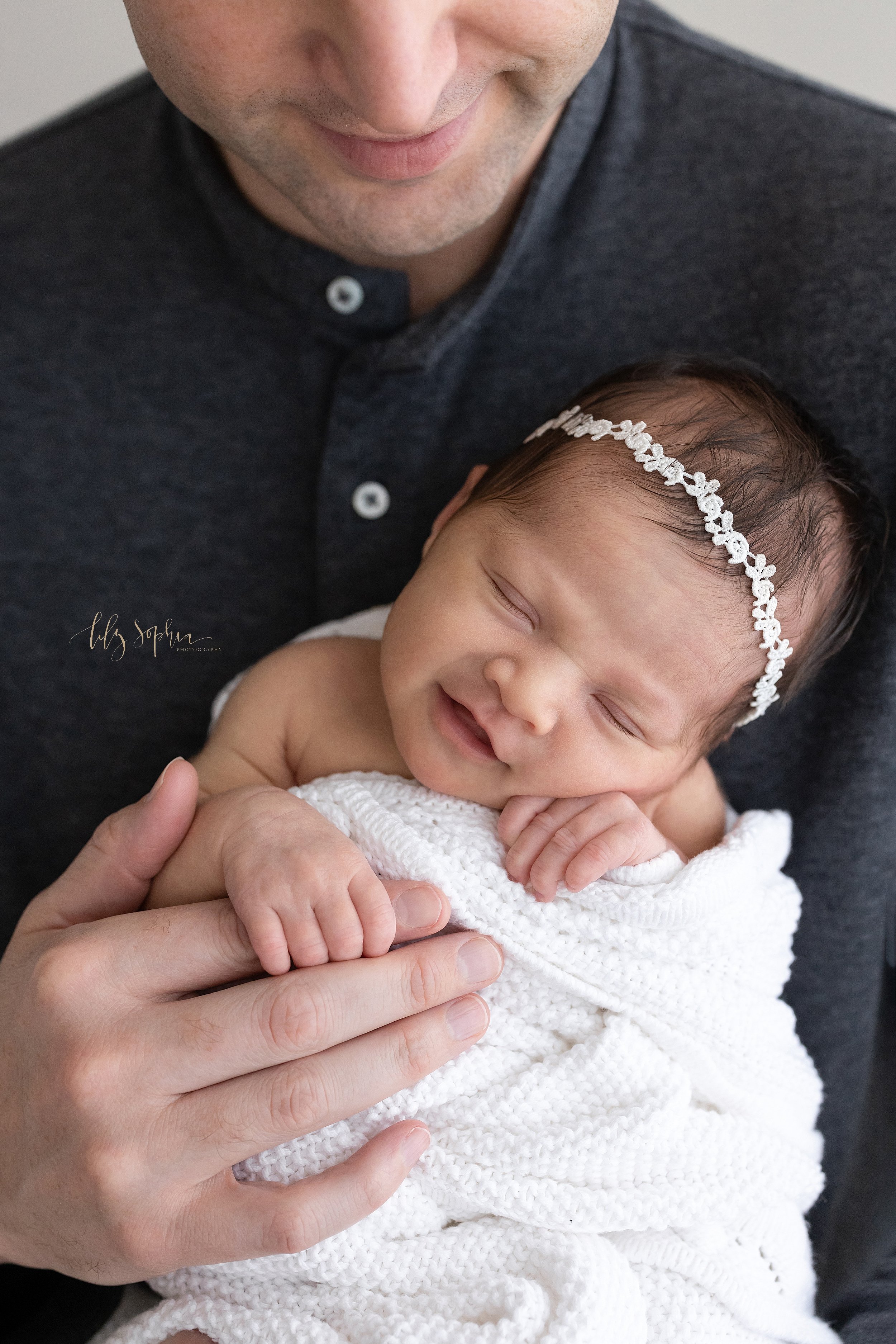  Close-up newborn photo of a smiling infant girl wearing a delicate headband in her wispy hair as she is held in front of her father’s chest and holds onto his index finger taken using natural light in a studio near Midtown in Atlanta, Georgia. 