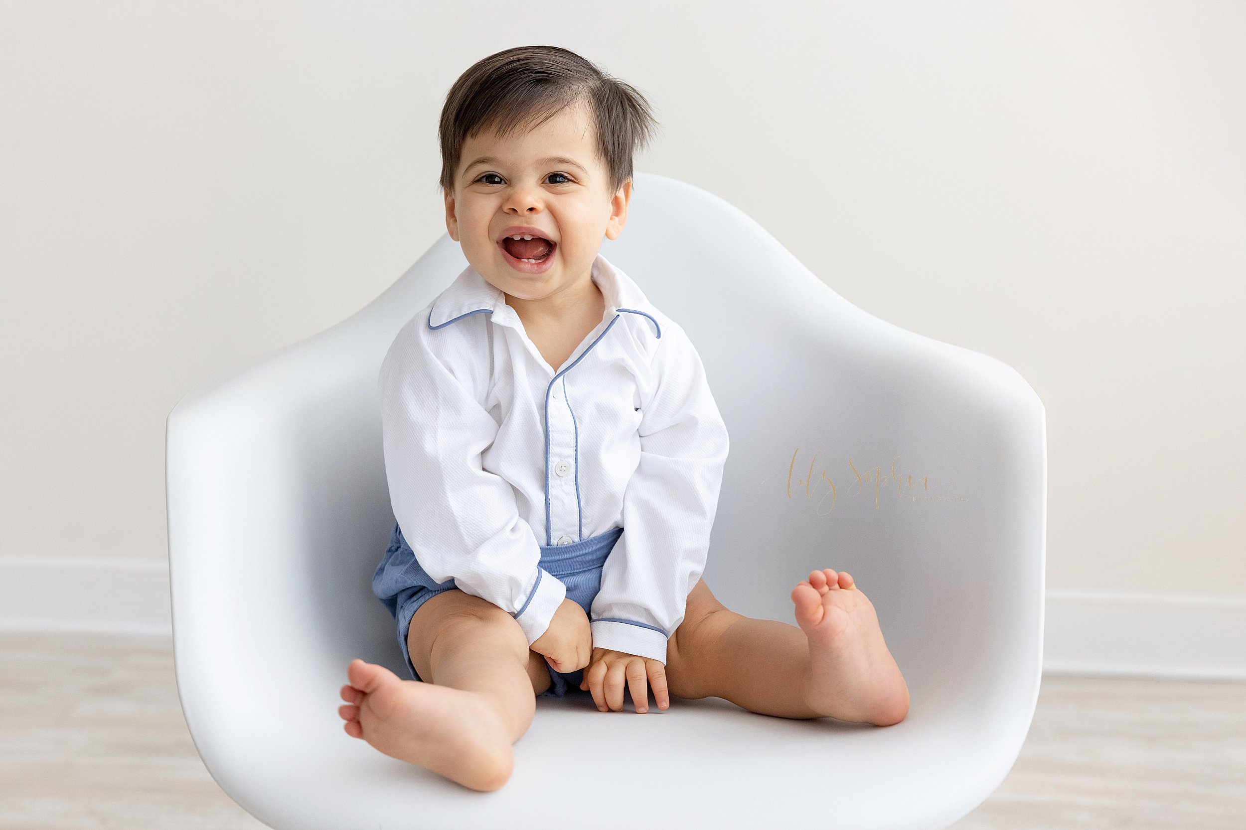  First birthday photo of a one year old little boy as he laughs while sitting in a white molded chair in front of a window streaming natural light in a studio near Old Fourth Ward in Atlanta. 