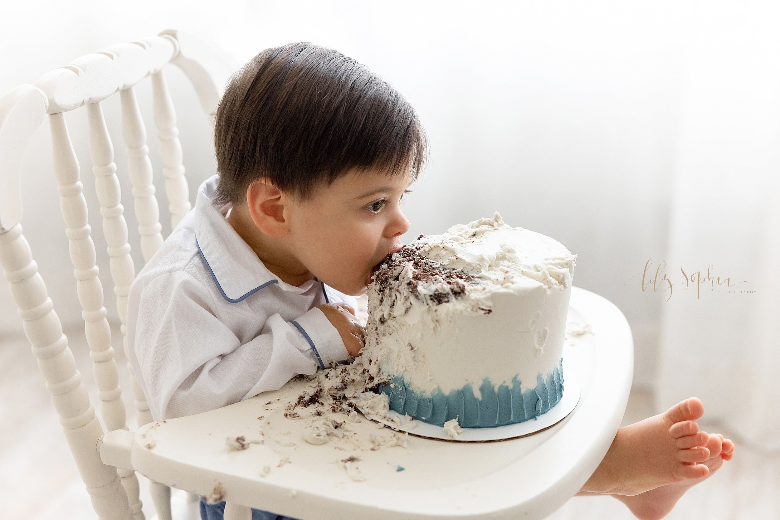  First birthday smash cake photo shoot with a one year old little boy seated in  an antique highchair with his smash cake on the tray in front of a window streaming natural light as he takes a bite out of the top of it taken near Virginia Highlands i