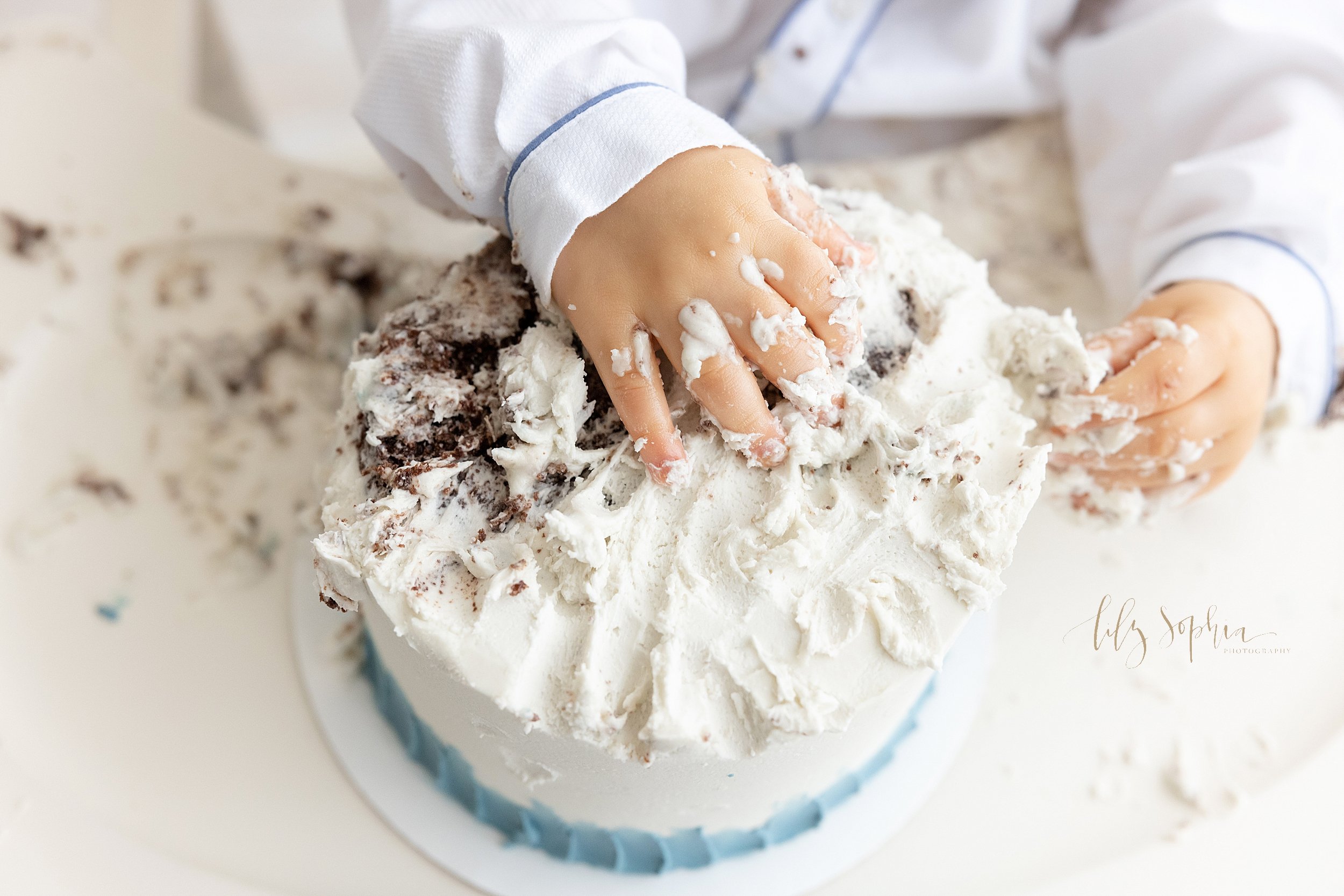  Cake smash picture of a one year old boy’s hands digging into his first birthday cake taken in a studio near Morningside in Atlanta that uses natural lighting. 