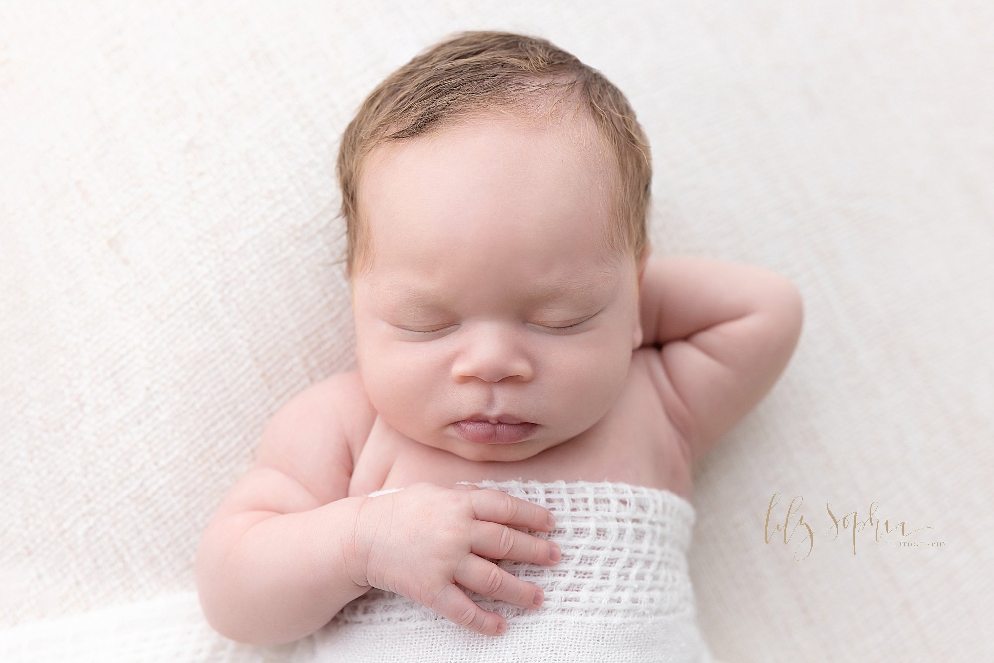  Newborn picture of a peacefully sleeping African-American baby boy with his left hand behind his head and his right hand resting on his chest while he lies on his back wrapped in a soft white knitted blanket taken in a natural light  studio near Dec