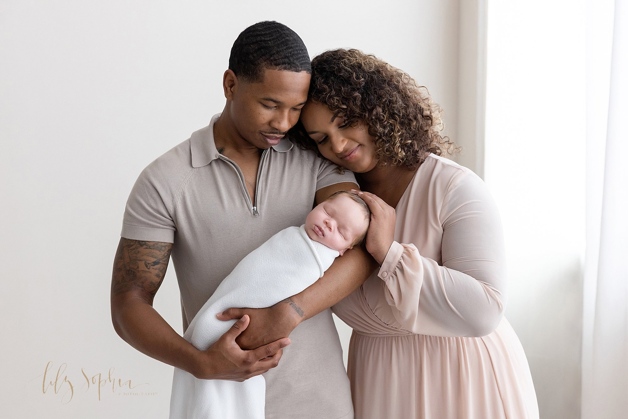  Family newborn photograph of an African-American father cradling his newborn son in his left arm as his wife stands next to him with her head on his shoulder and her left hand on top of her infant son’s head taken in front of a window streaming natu