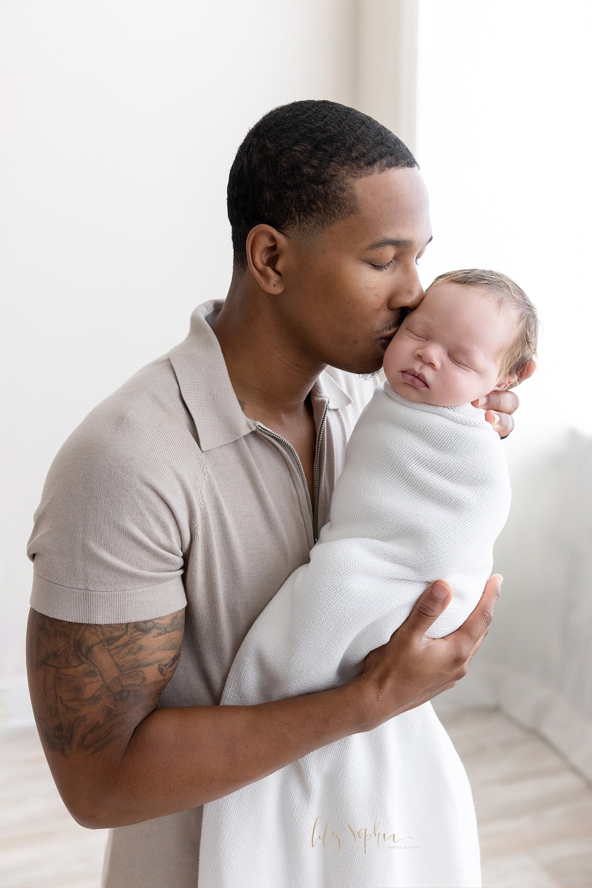 Newborn photo shoot with an African-American father holding his infant son in his hands in front of him as he kisses the cheek of his son as his son peacefully sleeps taken in front of a window streaming natural light near Oakhurst in Atlanta in a p
