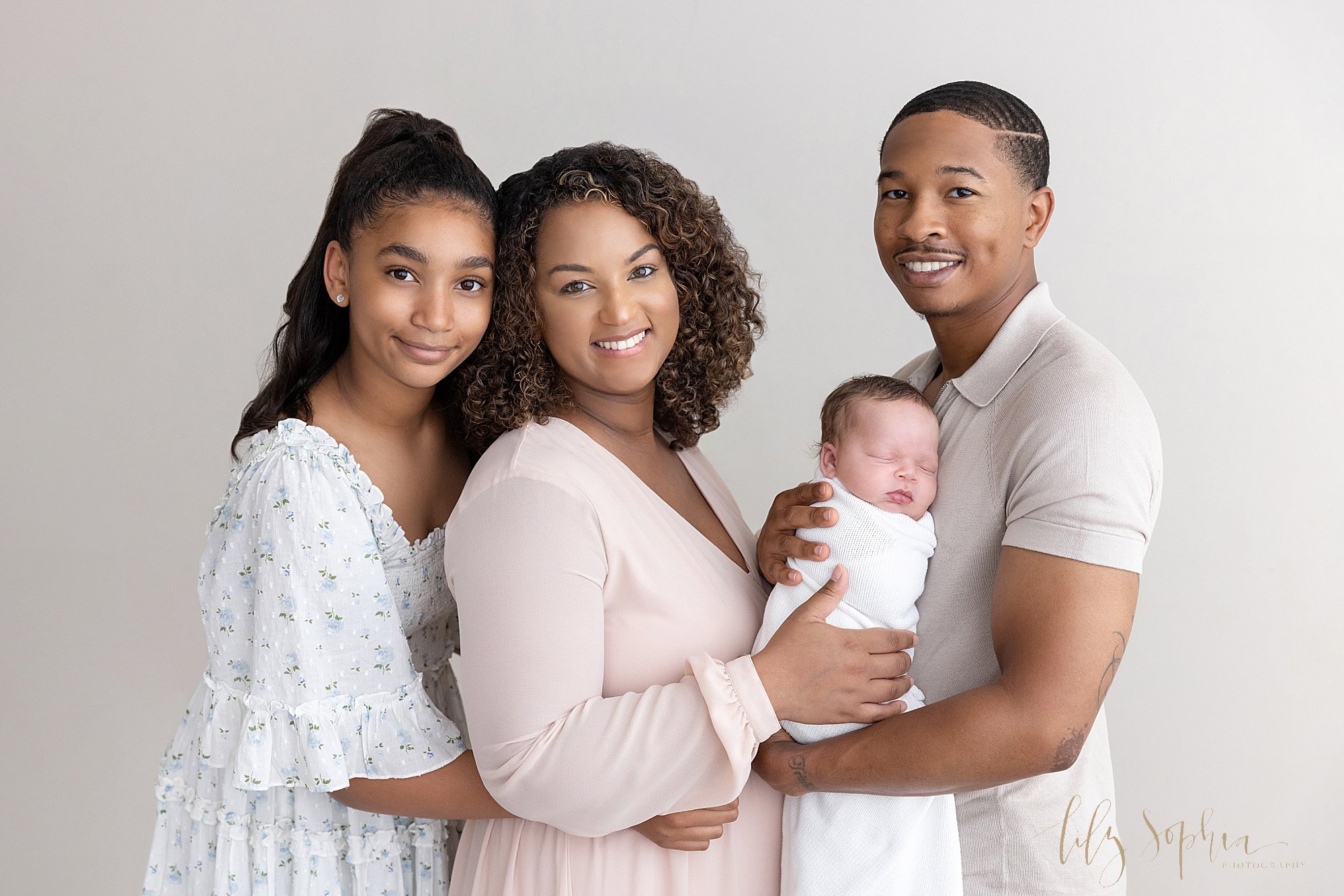  Family newborn photo session with an African-American dad holding his newborn son in front of him as he faces his wife who is standing in front of their teenage daughter taken near Kirkwood in Atlanta in a studio using natural light. 