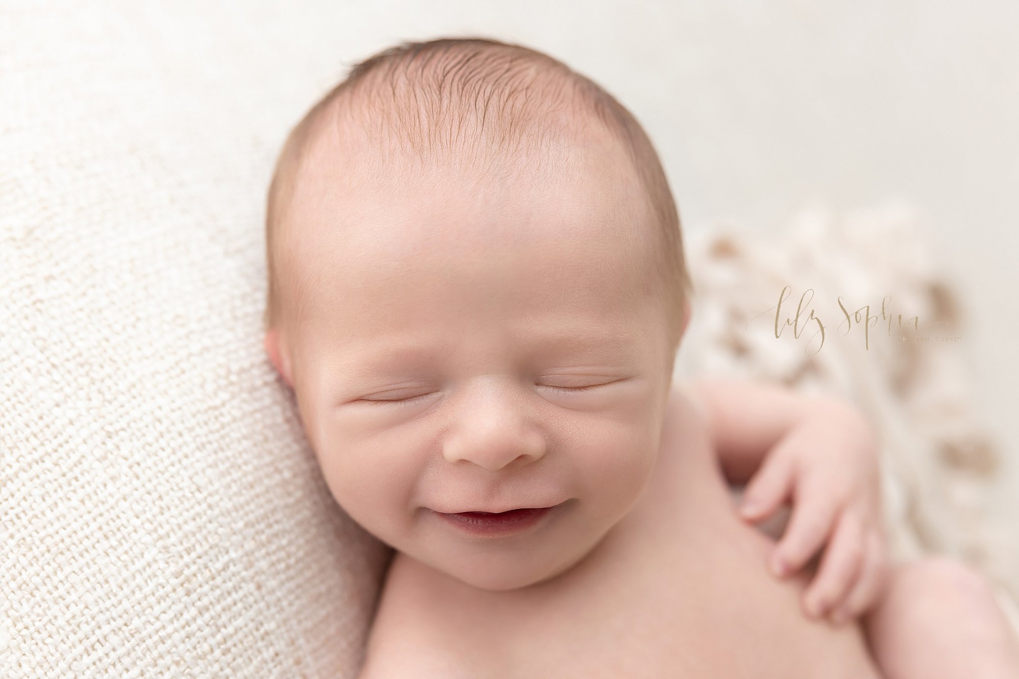  Newborn picture of a newborn baby boy as he smiles in his sleep while lying on his back with his head looking over his right shoulder taken in natural light in a photography studio located near Brookhaven in Atlanta, Georgia. 