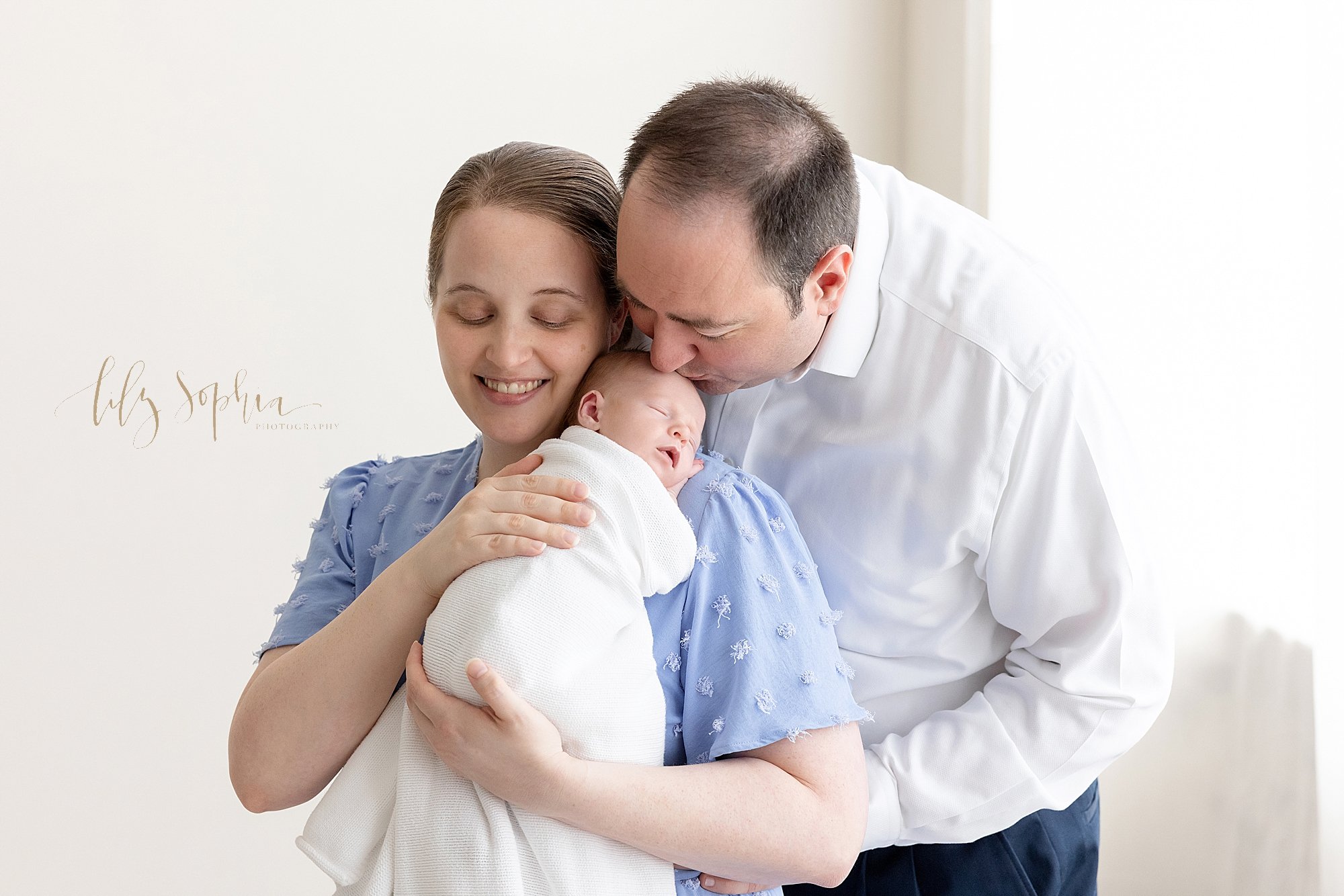  Newborn photo of a mother standing with her newborn baby son on her shoulder as her husband leans over her shoulder to kiss his son’s head as they stand in front of a window streaming natural light in a studio near Roswell in Atlanta, Georgia. 