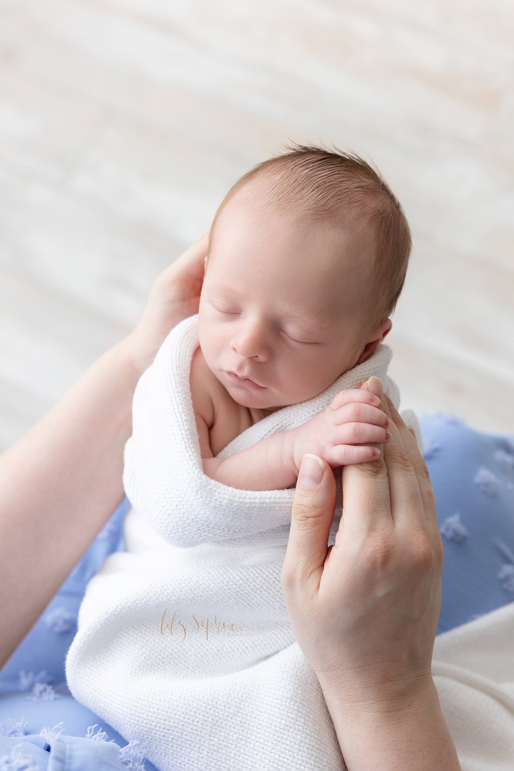  Newborn photo of a newborn baby boy wrapped in a soft white knitted blanket as his mother holds his head with her left hand and her son holds the index finger of her right hand taken in natural light near Vinings in Atlanta in a studio. 