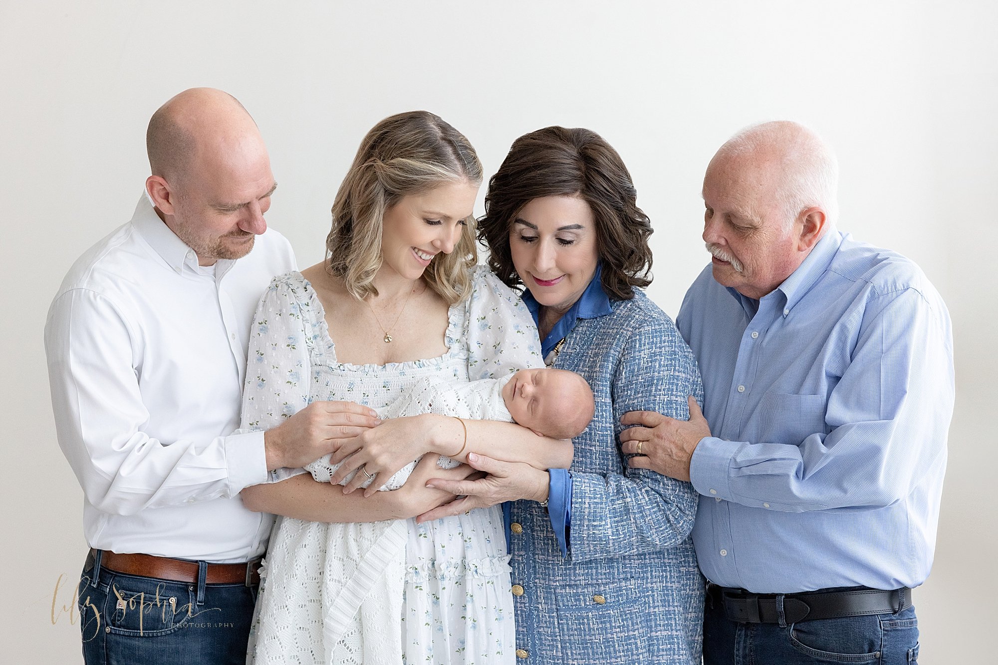 Newborn intergenerational picture of a mother cradling her newborn son in her arms with her husband stand to her left while her mother and father stand to her right as they all admire this new life in their family taken near Alpharetta in Atlanta in