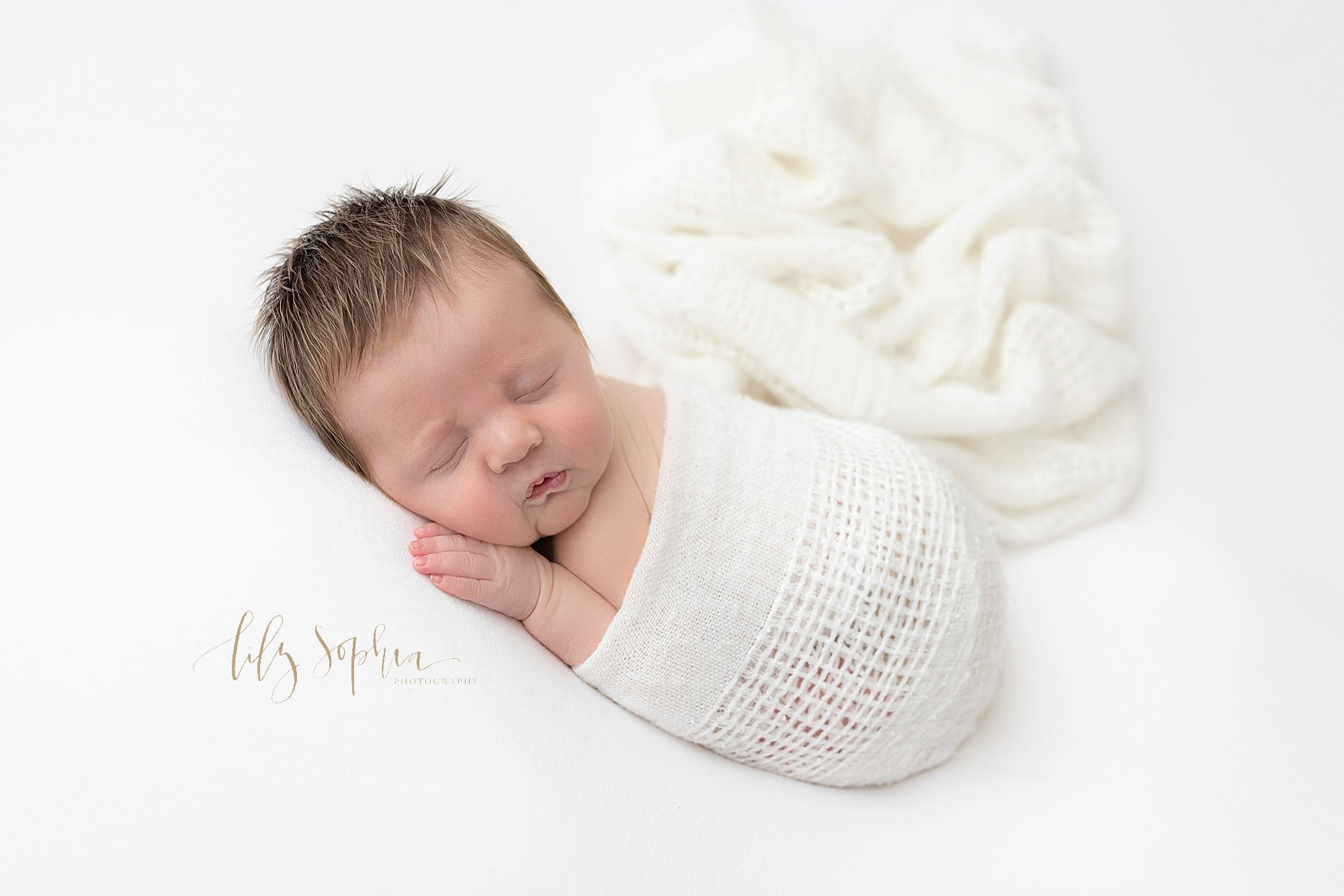  Newborn photo session of a newborn baby boy lying on his stomach with his hand under his right cheek as he is draped in a soft white knitted blanket taken in a natural light studio near Alpharetta in Atlanta, Georgia. 
