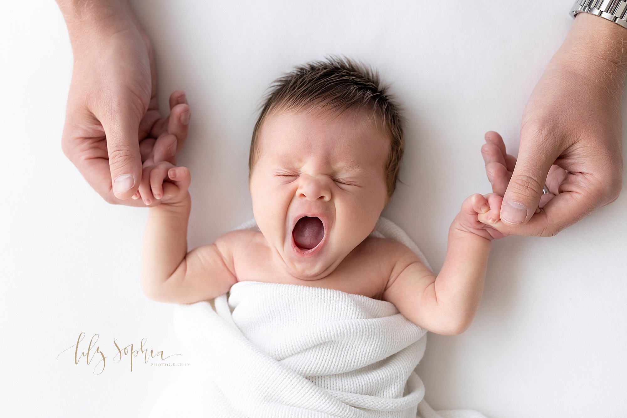  Sleepy newborn photo of a baby boy lying on his back wrapped in a white blanket as he holds his father’s index fingers and yawns taken near Ansley Park in Atlanta in a natural light studio. 