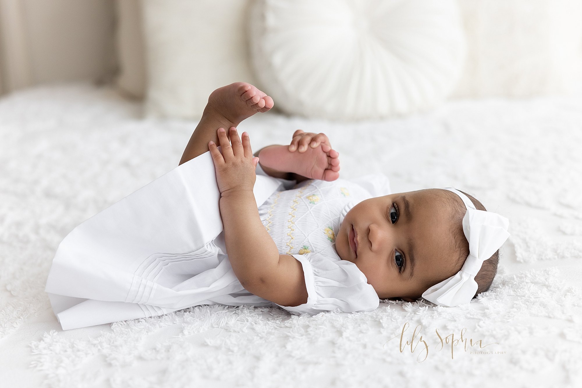  Milestone photo of an African-American six month old baby girl as she lies on her back wearing a white bow headband on her head and a white smocked short sleeved dress on top of a bed grabbing her foot with her right hand as she looks over her left 