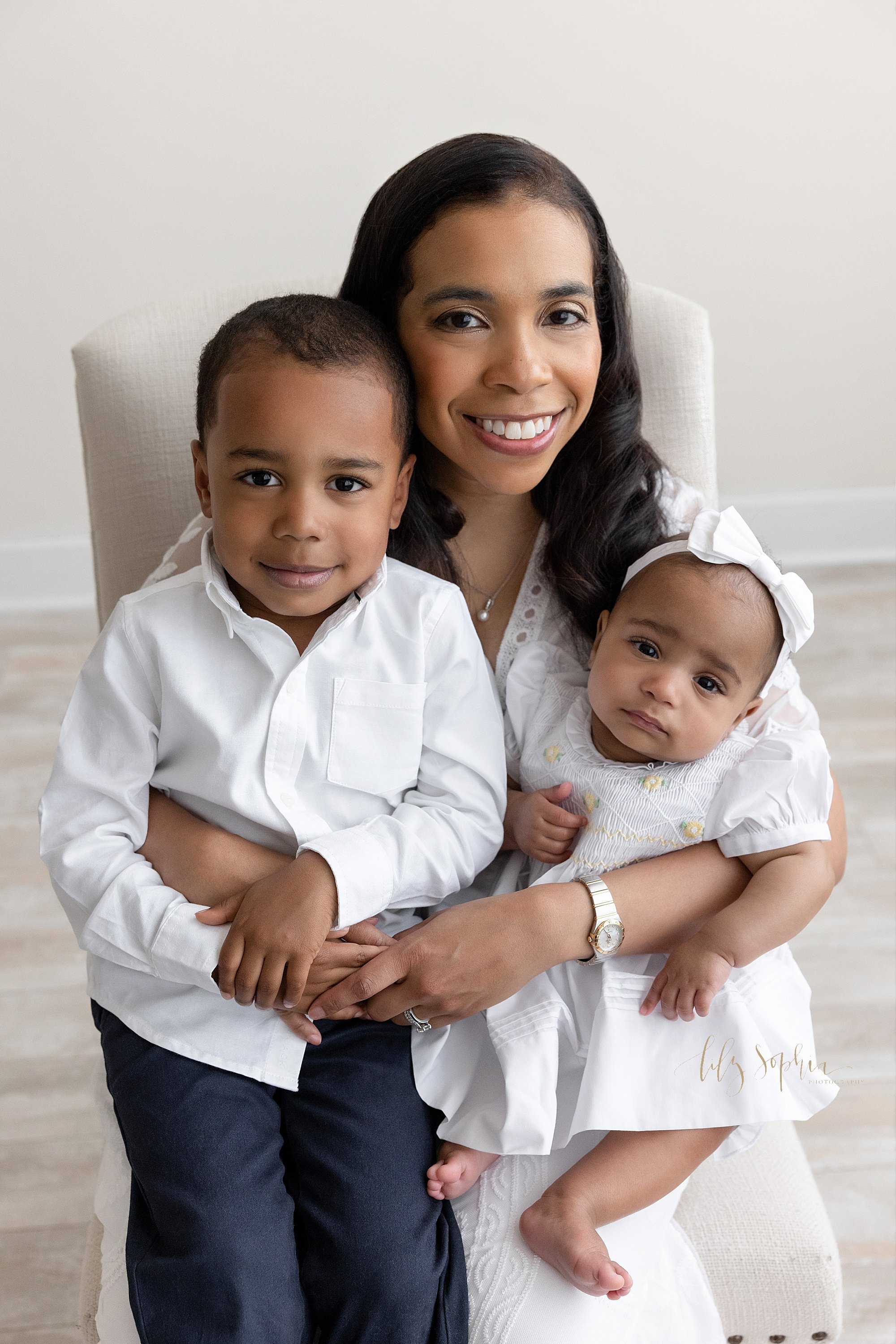  Family photo of an African-American mother sitting in an upholstered chair with her six month old baby girl on her left knee and her young son on her right knee taken near Morningside in Atlanta in natural light in a photography studio. 