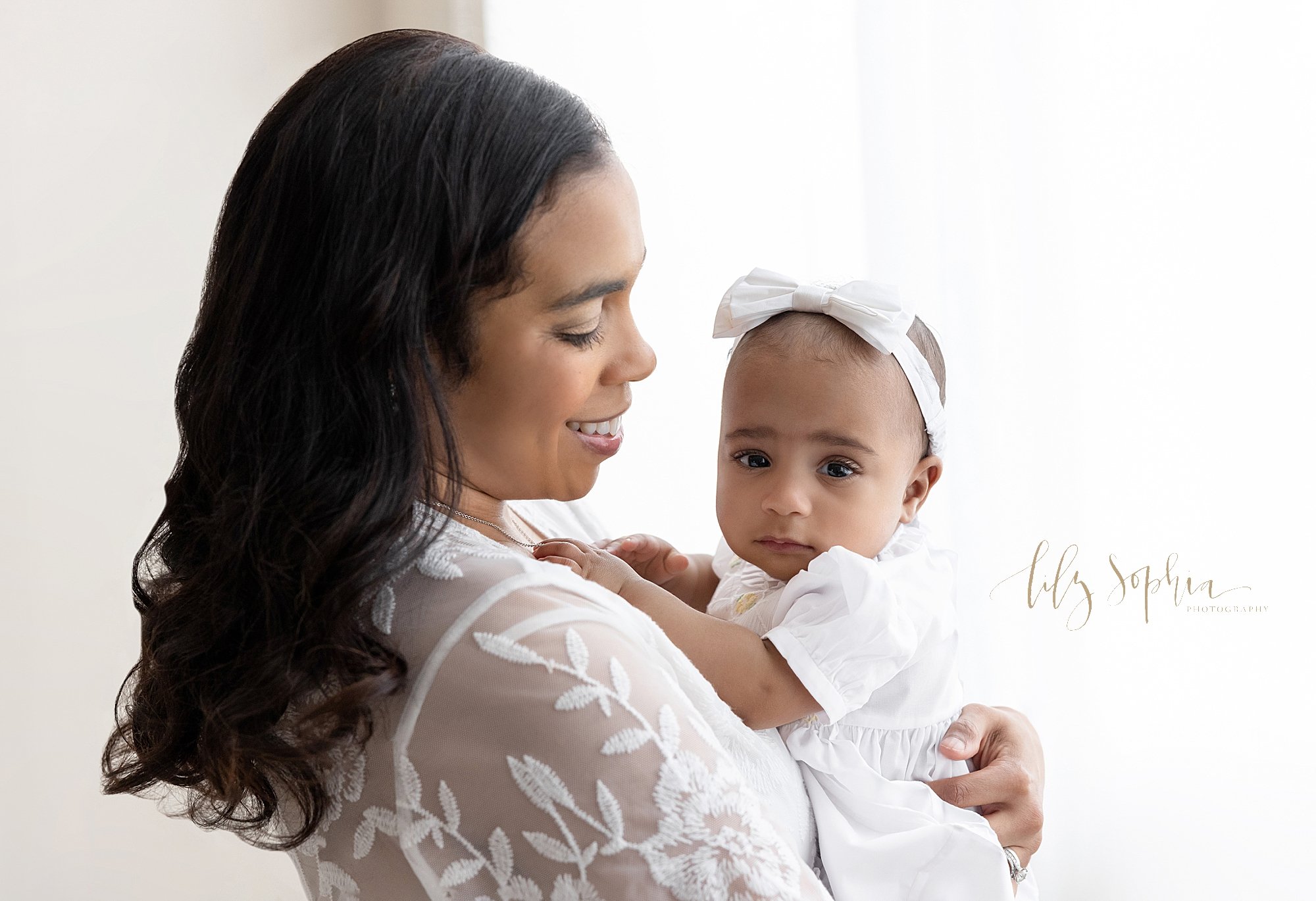  Milestone baby photo of a six month old African-American baby girl as she is held in her mother’s arms wearing a bow headband on her head as mom stands in front of a window streaming natural light in a studio near Virginia Highlands in Atlanta. 