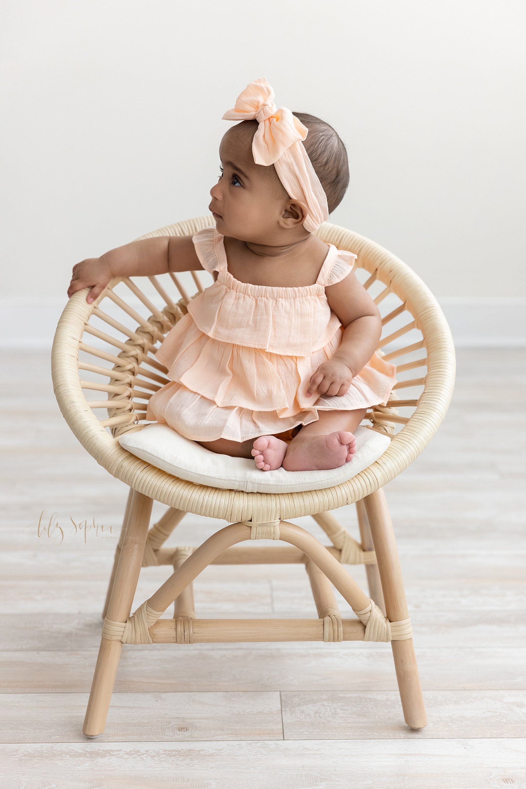  Profile milestone portrait of a six month old African-American baby girl as she sits in a rattan chair wearing a peach  ruffled sundress with a bow headband on her head taken near Brookhaven in Atlanta in a natural light studio. 