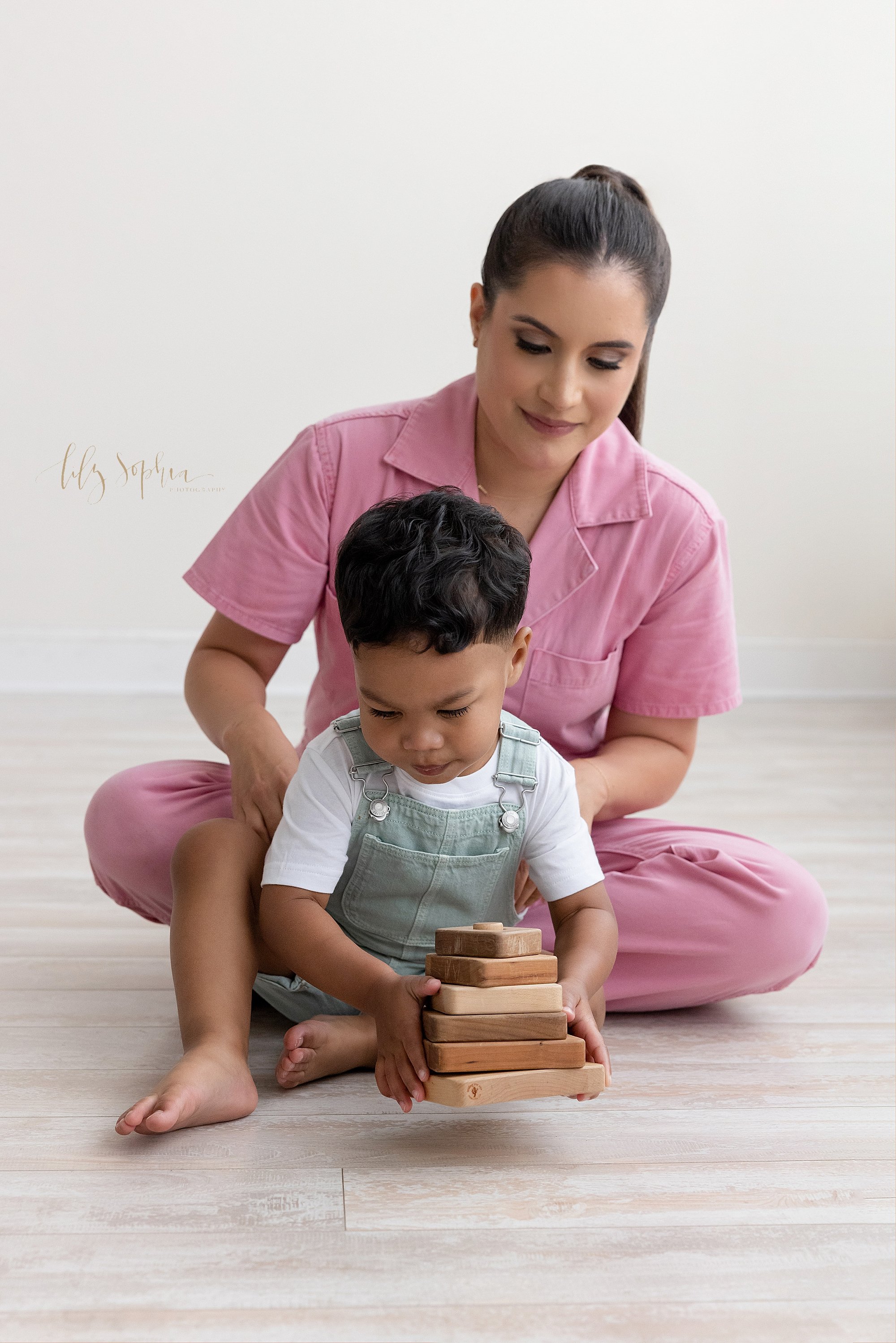  Family picture of a mother sitting behind her toddler son as he plays with a wooden stacking toy on the floor of a photography studio next to a window streaming natural light located near Sandy Springs in Atlanta, Georgia. 