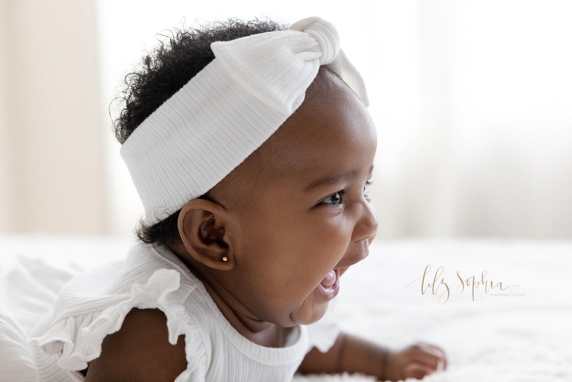  Close-up baby portrait of the profile of an laughing African-American baby girl as she lies on her stomach on top of a bed wearing a white dress and a white headband with a bow on her head in front of a window streaming natural light in a photograph