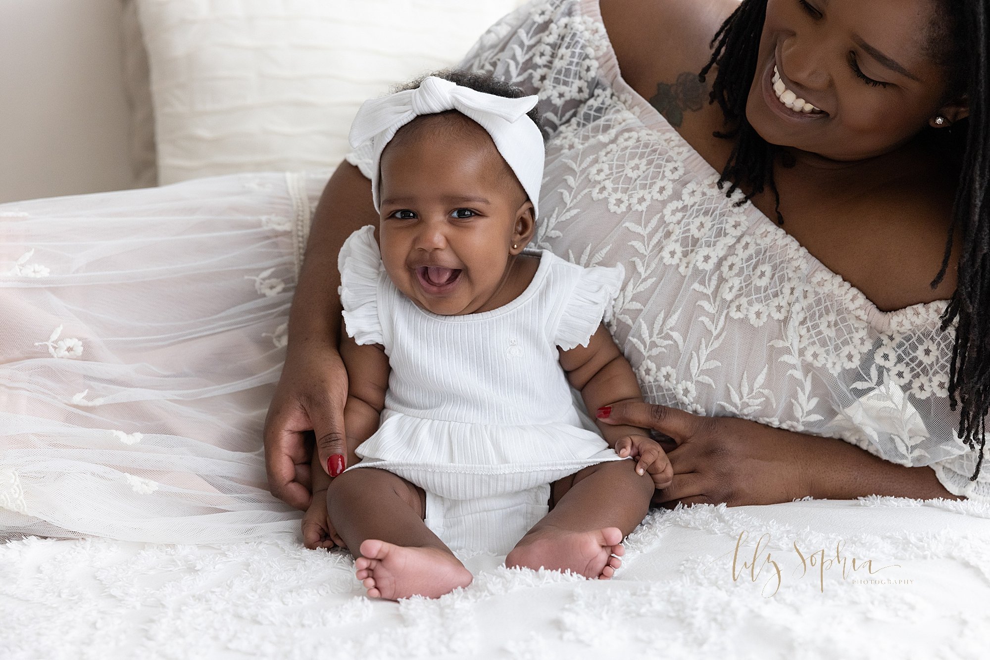  Baby photo shoot of an African-American baby girl as she sits on top of a bed with her mom leaning on her right arm as she lies on the bed behind her taken using natural light in a studio near Oakhurst in Atlanta. 