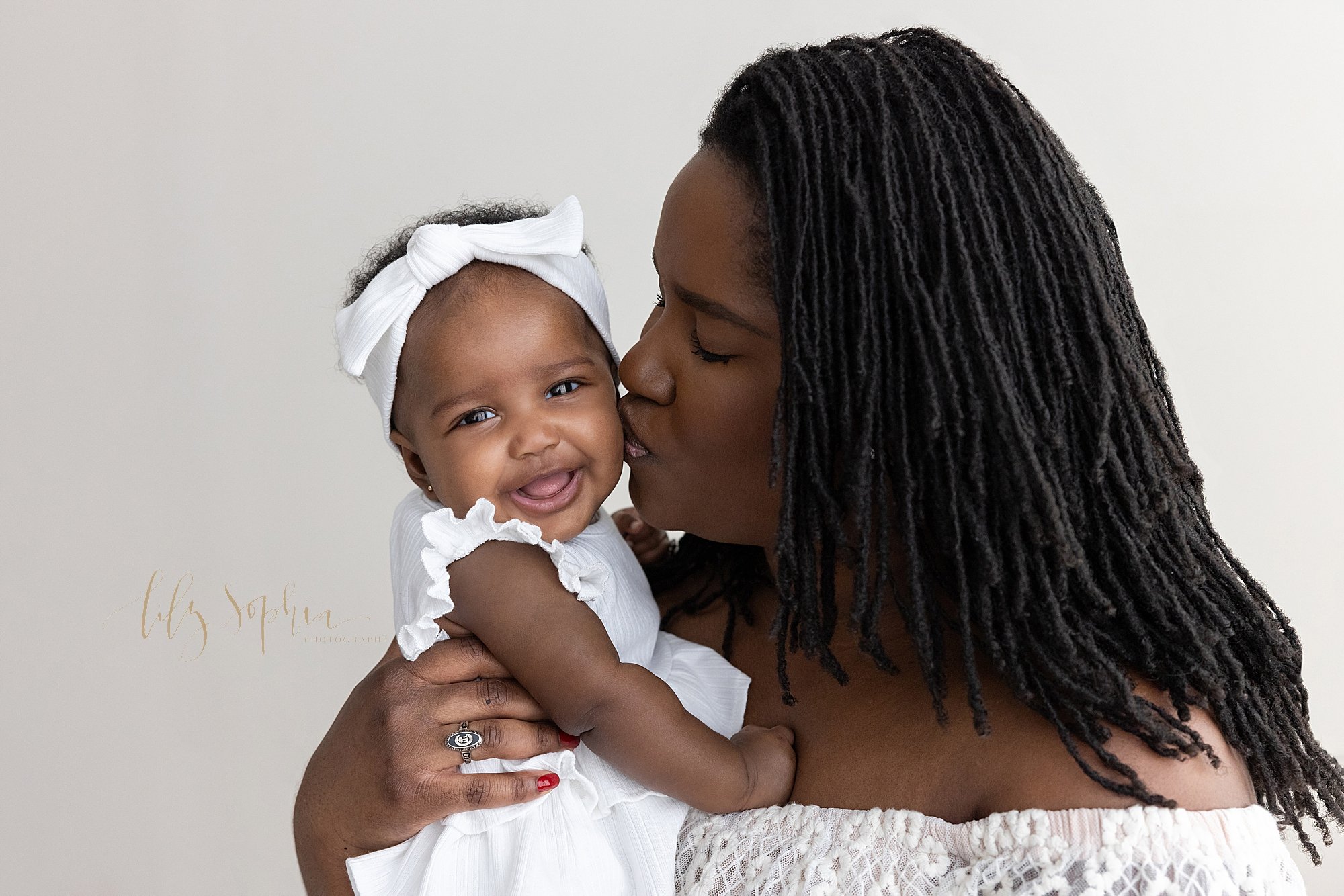  Baby photo session with a smiling African-American baby girl dressed in white and wearing a white headband in her hair as her mother holds her and kisses her cheek taken near Old Fourth Ward in Atlanta in a natural light studio. 