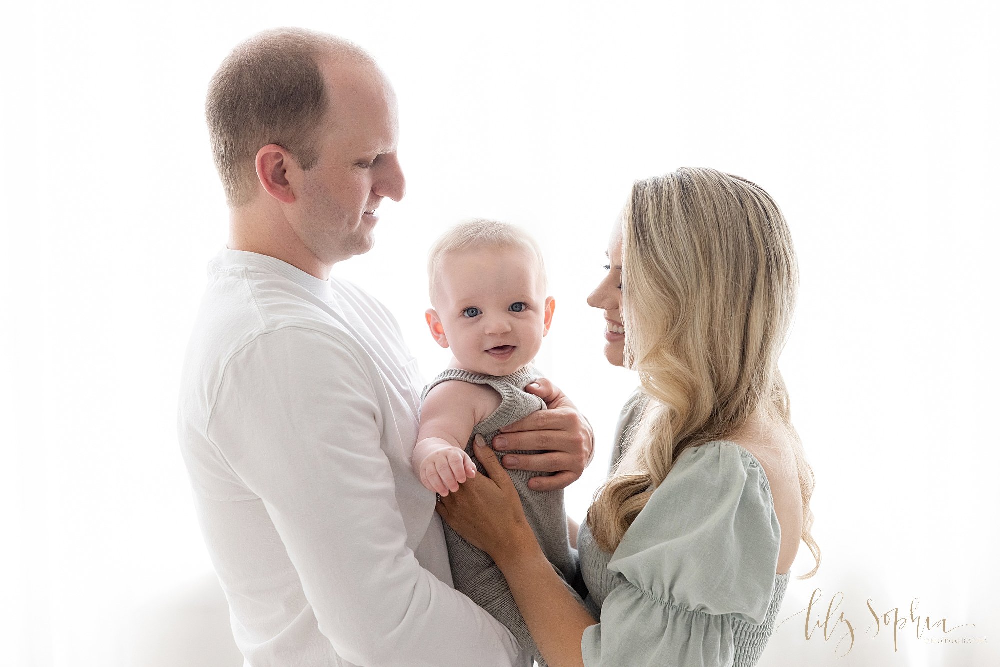  Family photo session of a father holding his baby boy in front of him as his wife faces him to take him into her arms as the the couple stands in front of a window streaming natural light in a studio near Roswell in Atlanta, Georgia. 