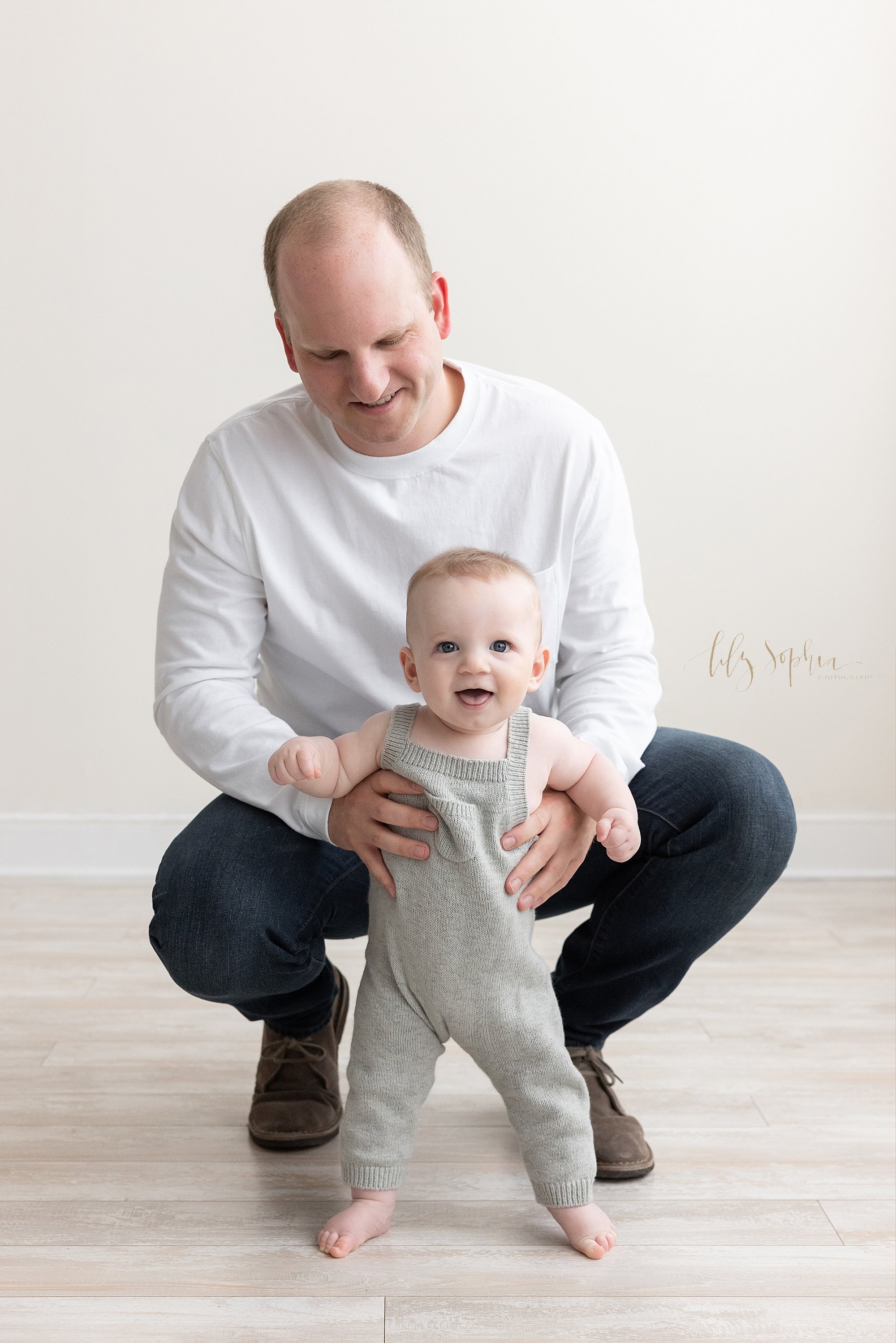  Family photo session of a father squatting behind his baby boy as he supports the son who is trying to stand in a studio near Buckhead in Atlanta that uses natural light. 