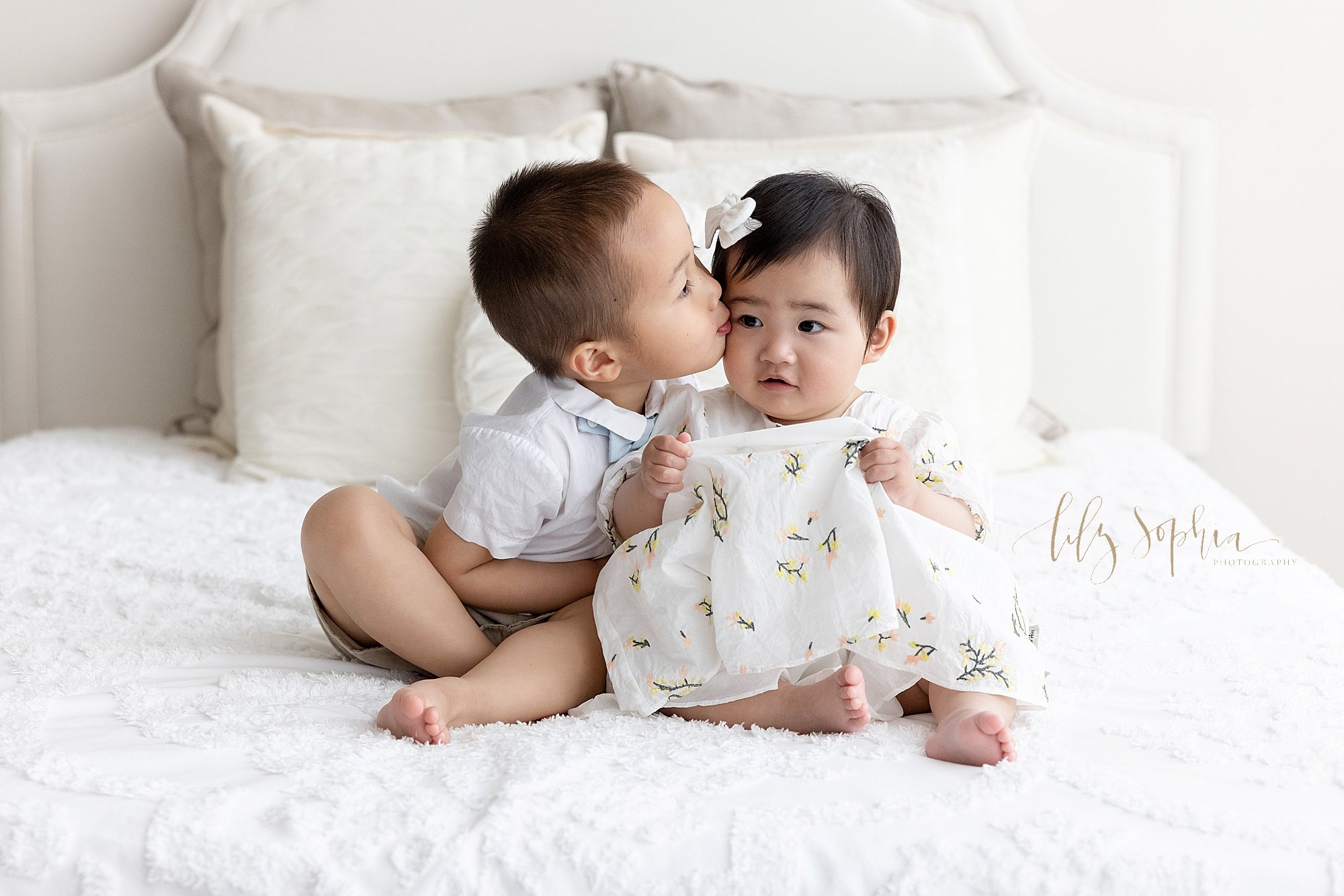  Family portrait of sibling love as an Asian toddler brother kisses his one year old sister as they sit atop a bed next to a window streaming natural light in a studio near Buckhead in Atlanta. 